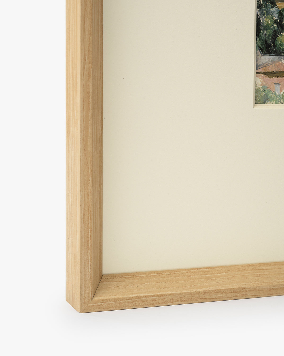 Close-up of a museum-quality natural woodgrain frame with cream matting, showing a small section of Le Village artwork in the upper right corner on a white background, by Vendixs