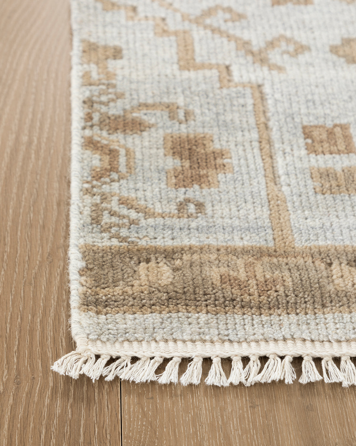 A close-up of the Lexington Hand-Knotted Wool Rug shows its geometric beige and light brown pattern on a wooden floor, finished with short white fringe along the edge, by Vendixs