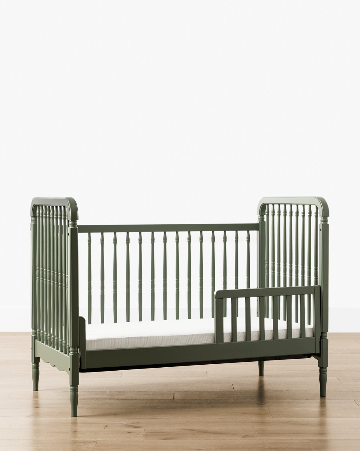 The Liberty 3-in-1 Convertible Spindle Crib with Toddler Bed Conversion Kit, crafted from solid pine and featuring slatted sides plus a partial safety rail, is shown on a wooden floor against a white background, by Vendixs