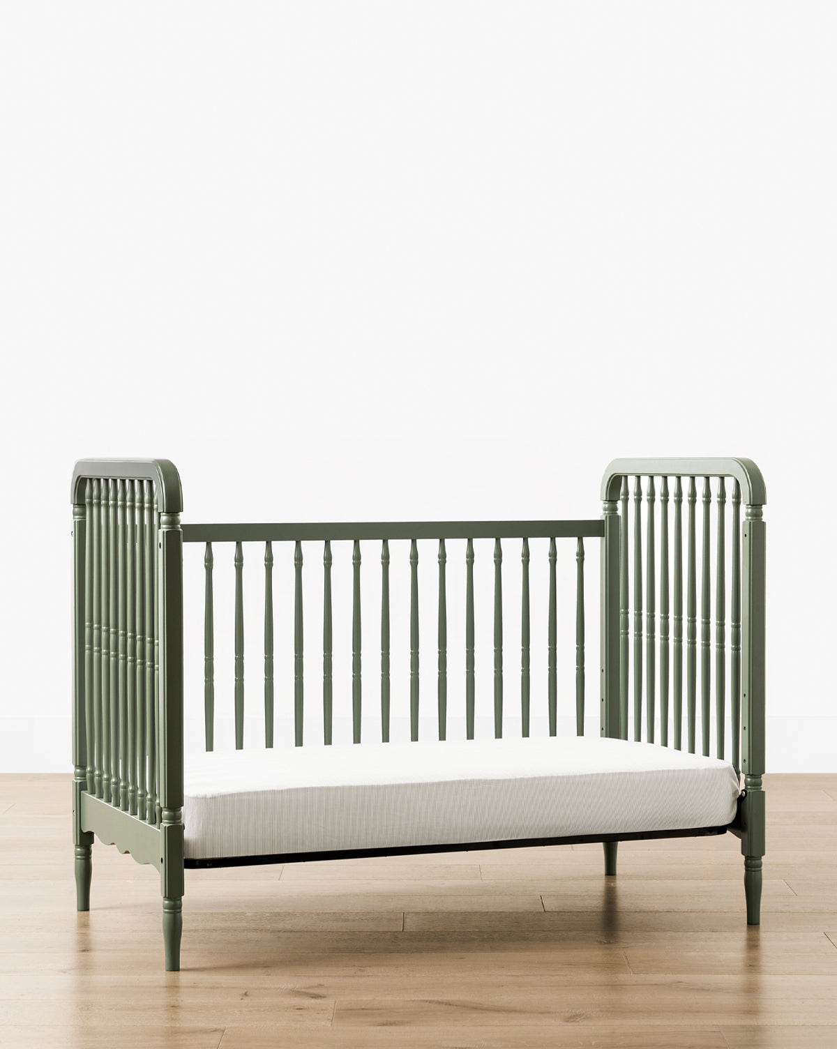 The Liberty 3-in-1 Convertible Spindle Crib with Toddler Bed Conversion Kit, shown in green solid pine with slatted sides and a white mattress, stands on a light wood floor against a plain white background by Vendixs