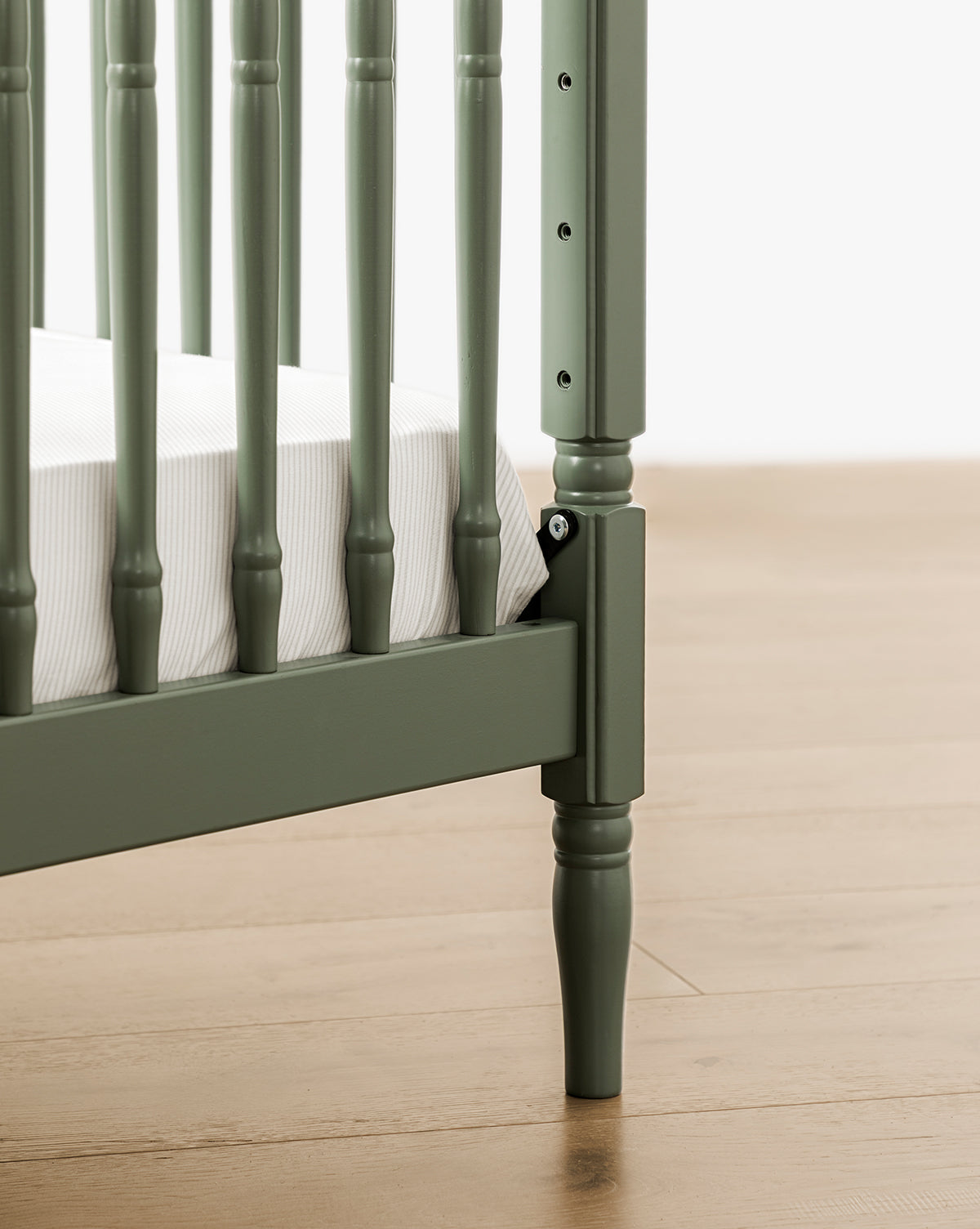 Close-up of the Liberty 3-in-1 Convertible Spindle Crib with Toddler Bed Conversion Kit in green solid pine, featuring vertical slats, a white mattress, and light wood flooring in the background, by Vendixs