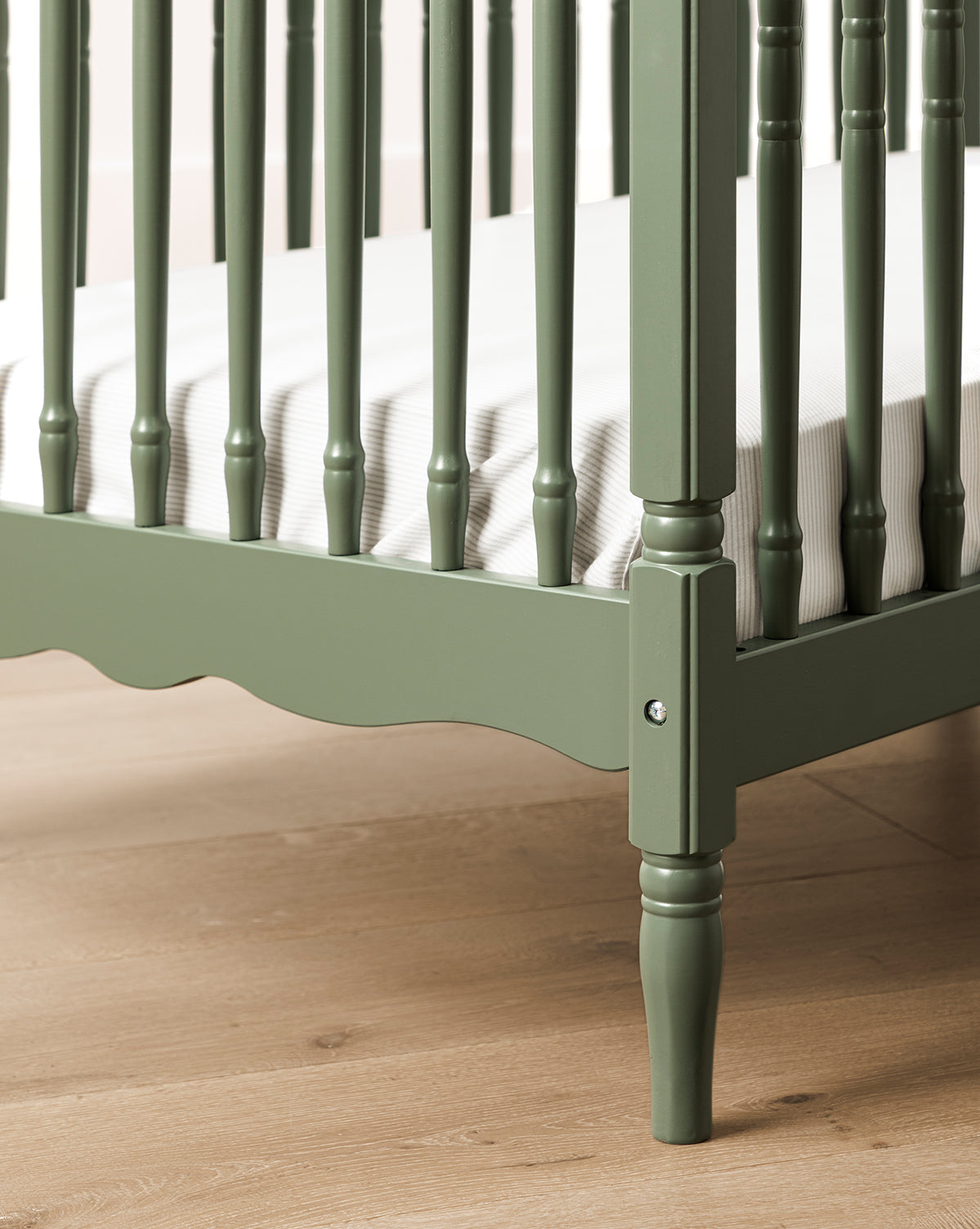 A close-up of the Liberty 3-in-1 Convertible Spindle Crib w/Toddler Bed Conversion Kit in green, crafted from solid pine with vertical slats, decorative spindles, a white striped mattress, and set on a light wooden floor, by Vendixs