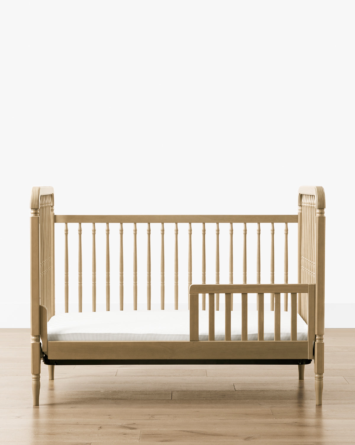 The Liberty 3-in-1 Convertible Spindle Crib with Toddler Bed Conversion Kit, crafted from solid pine, features spindle rails and an open side with a safety guard. It sits on a light wood floor against a plain white background, by Vendixs
