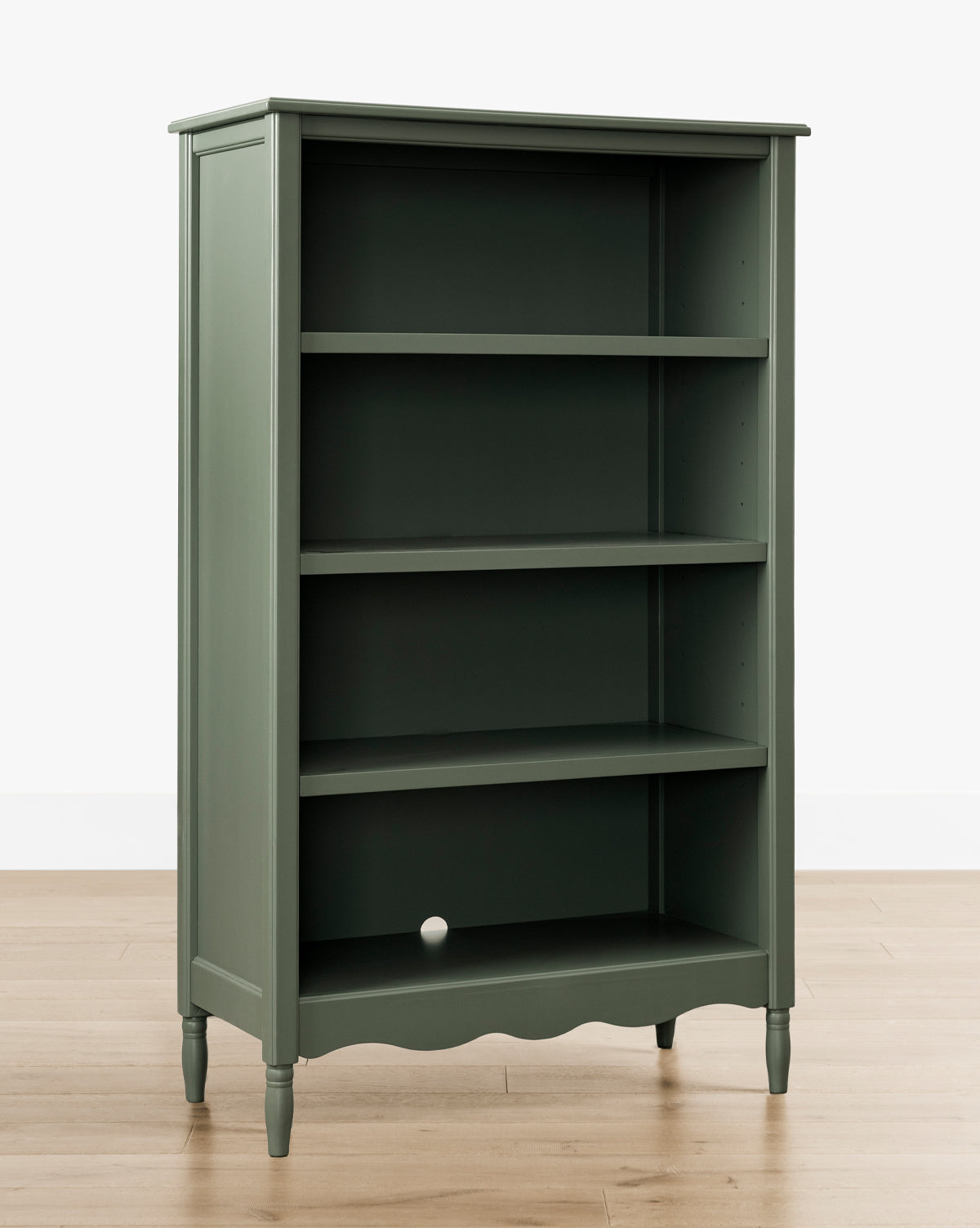 The Liberty Bookcase in green, featuring four shelves, is displayed against a white wall on a light wood floor, highlighting its heirloom craftsmanship. A small white ball rests on the bottom shelf, by Vendixs