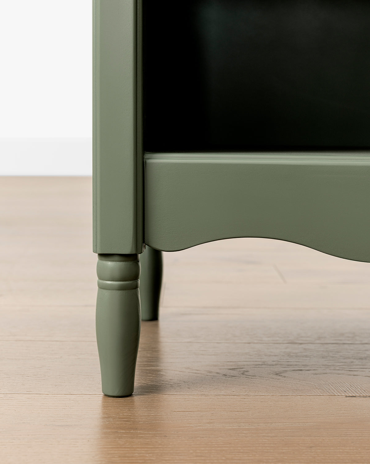 Close-up of the bottom corner of the green Liberty Bookcase with turned legs, highlighting heirloom craftsmanship, on a light wood floor against a white wall, by Vendixs