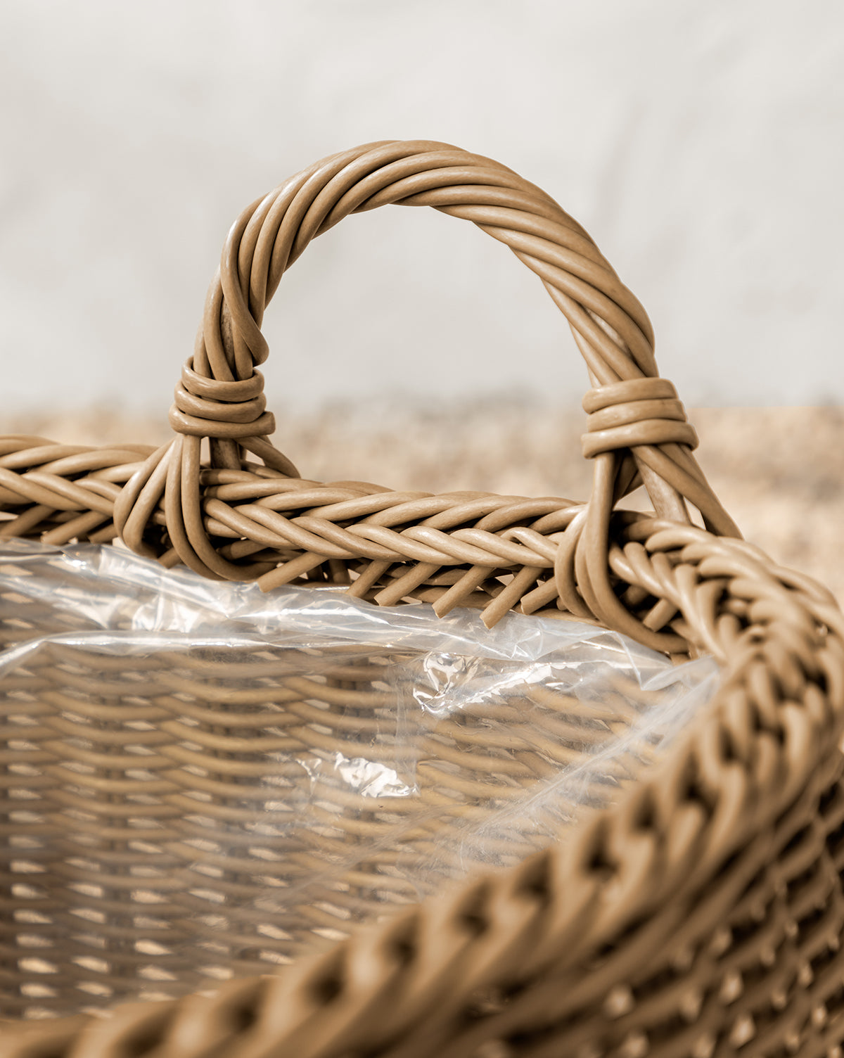 The Lightwash Handled Planter Basket features a sturdy twisted handle and a clear plastic liner, making it perfect for indoor plants. Its poly-rattan texture stands out against a softly blurred, neutral background, by Vendixs