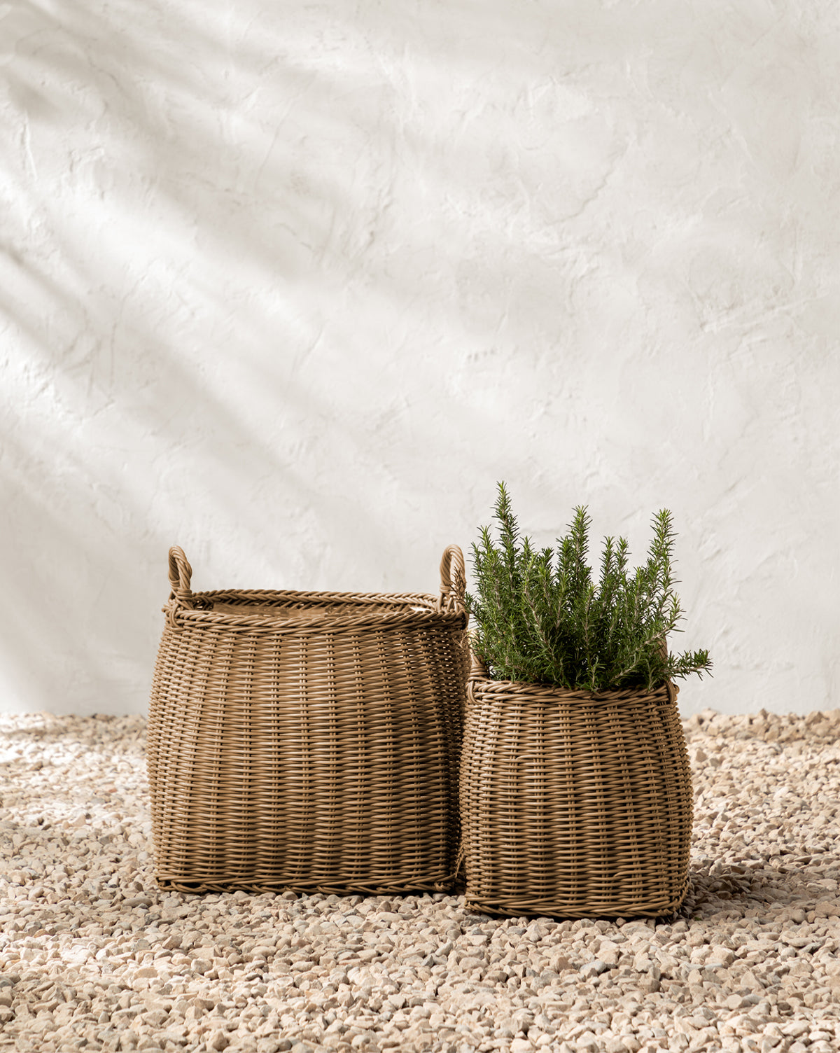 Two Makerspalm Lightwash Handled Planter Baskets rest on pebbled ground; the larger is empty with handles, and the smaller holds a green rosemary plant. Both sit by a textured white wall in soft natural light, Vendixs