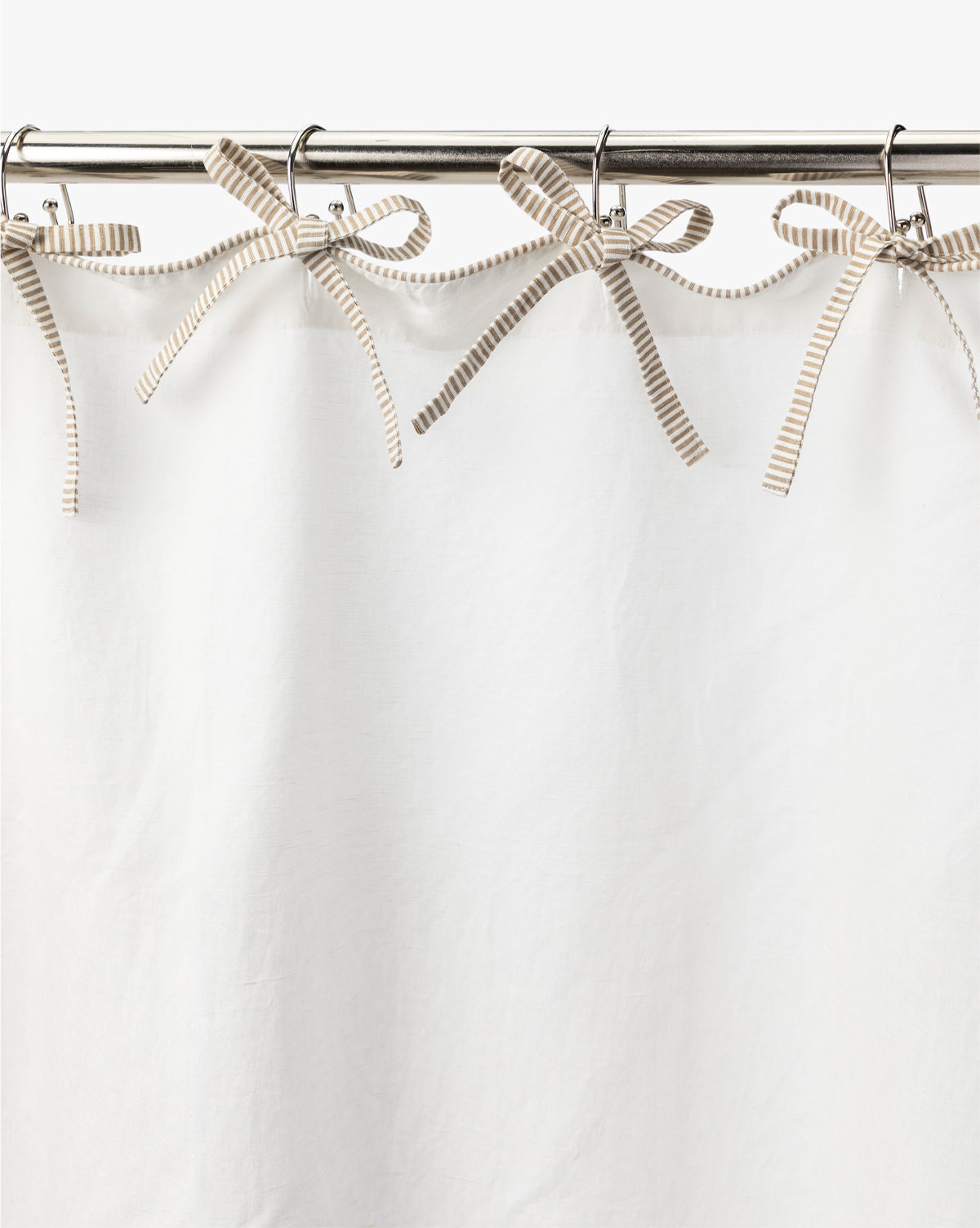 The Lillian Shower Curtain with Striped Ties is a white linen curtain that hangs from a metal rod, secured at the top with decorative beige and white striped ribbon ties fashioned into bows, by Vendixs