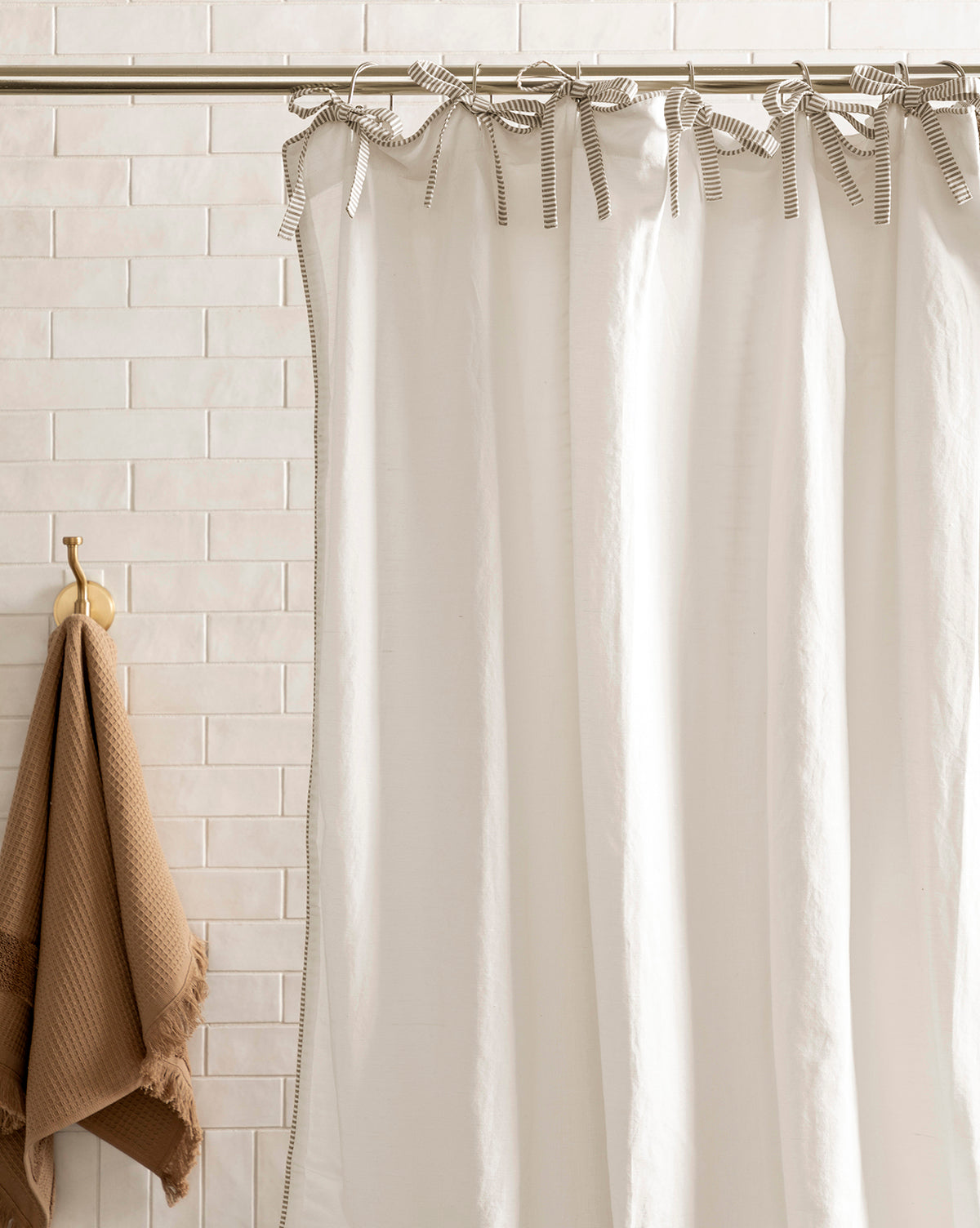 The Lillian Shower Curtain with Striped Ties hangs from a rod in front of light beige tiles, while a brown textured towel is displayed on a gold hook to the left, by Vendixs