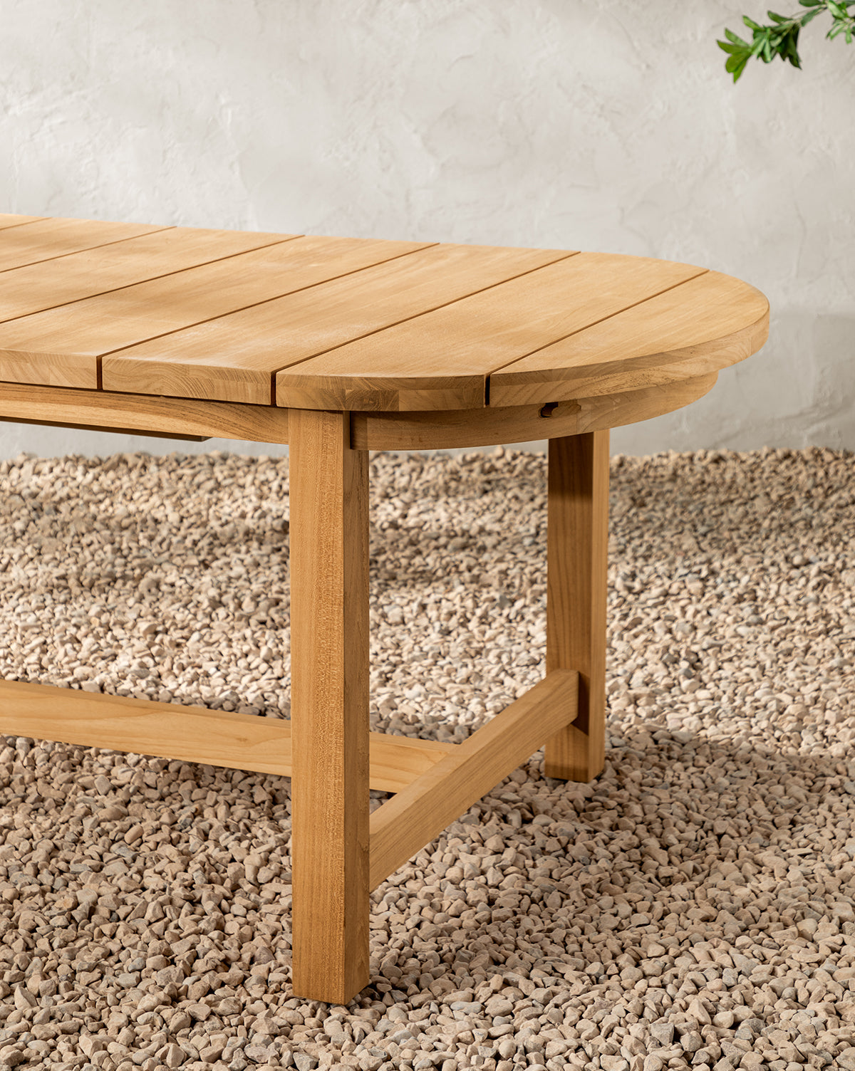 The Linwood Teak Extension Dining Table features a slatted oval top and sturdy legs, shown here in close-up on pebbles against a light textured wall, by Vendixs