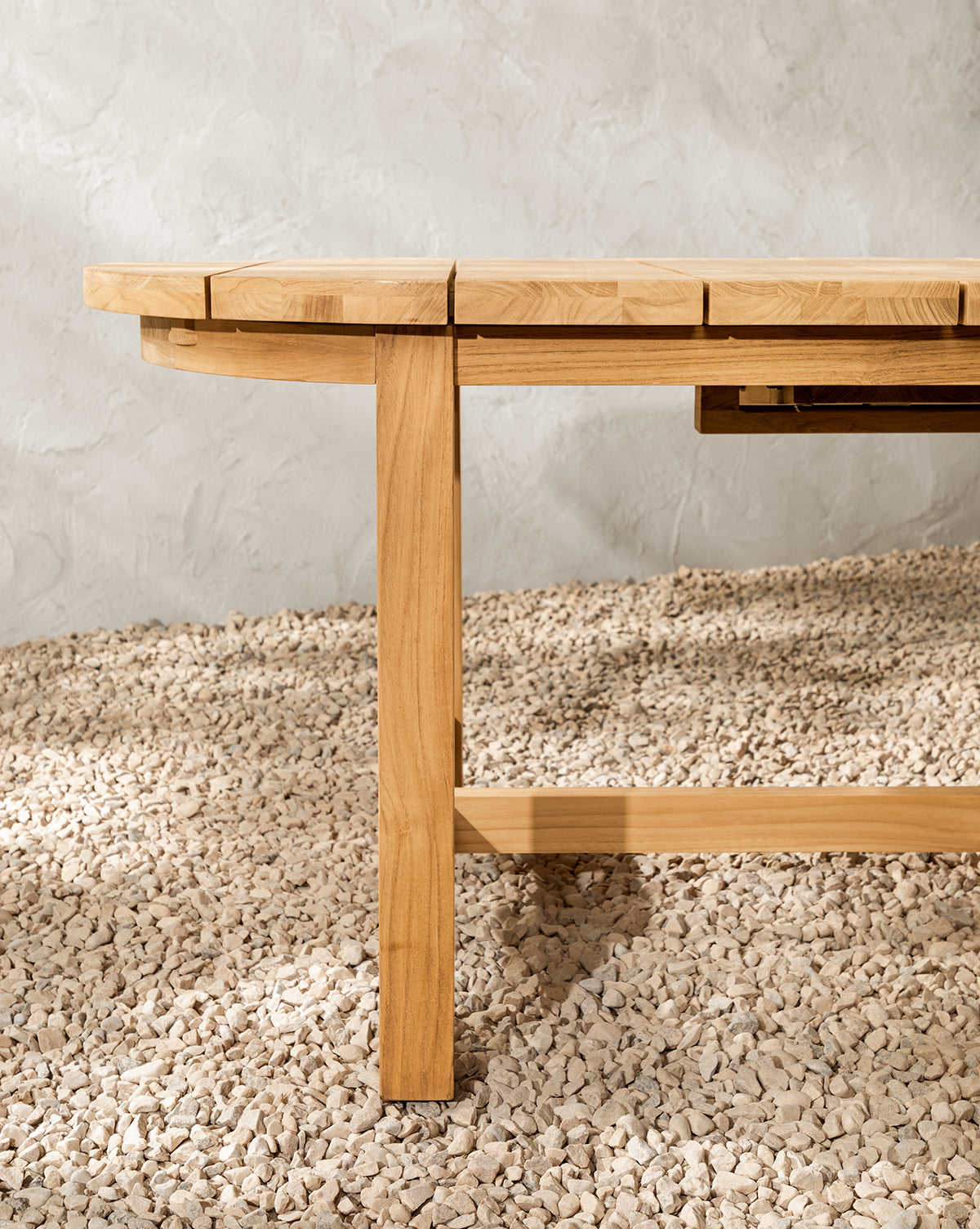 A close-up of the Linwood Teak Extension Dining Table leg and edge, crafted from light wood, shown on a textured, light-colored pebble floor with a gray textured wall in the background, by Vendixs