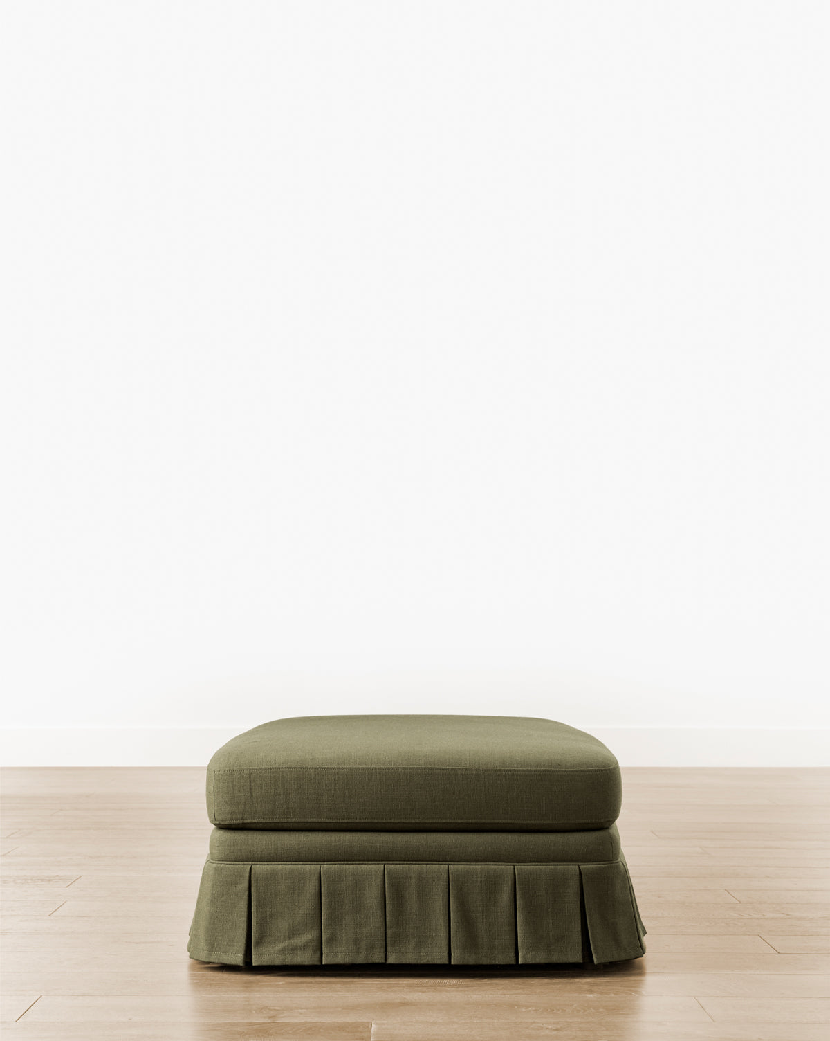 The Louisa Ottoman showcases custom green upholstery and a tailored box pleat skirt, elegantly placed on a light wood floor with a simple white wall backdrop, by Vendixs