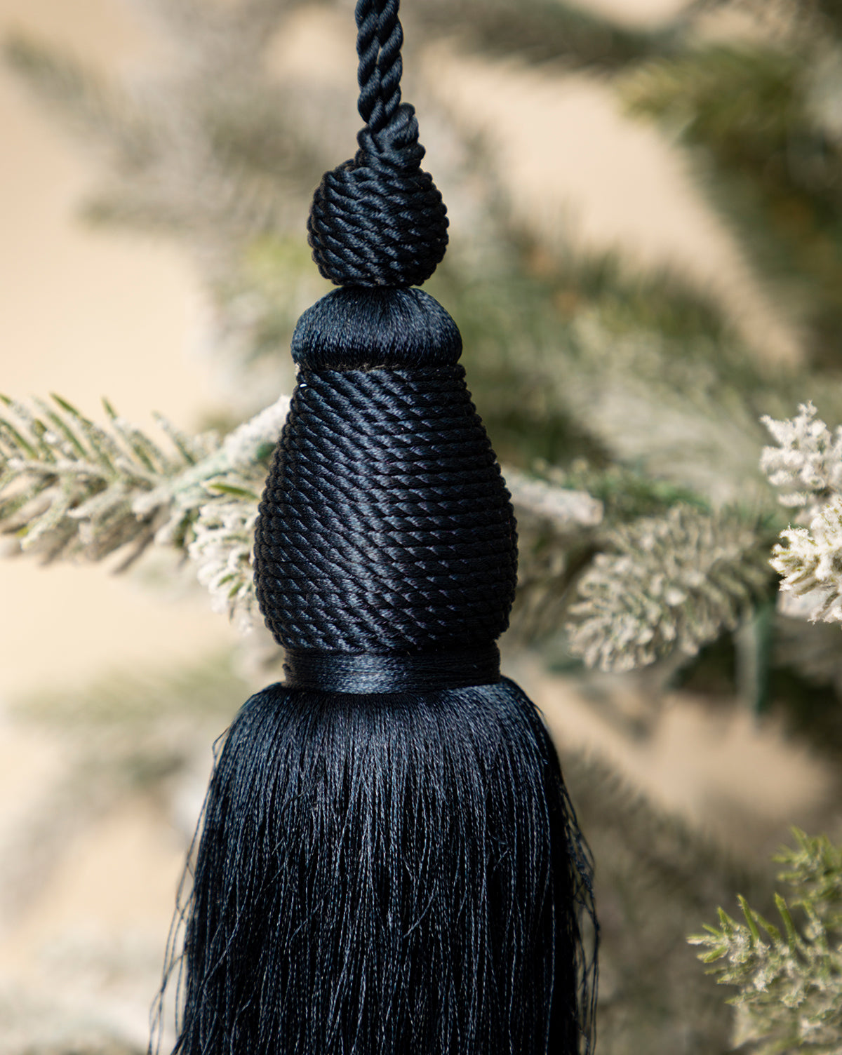 A close-up of the Lucia Bullion Tassel, a black vintage-inspired holiday ornament with silky threads, hanging on a snow-dusted artificial pine branch, by Vendixs
