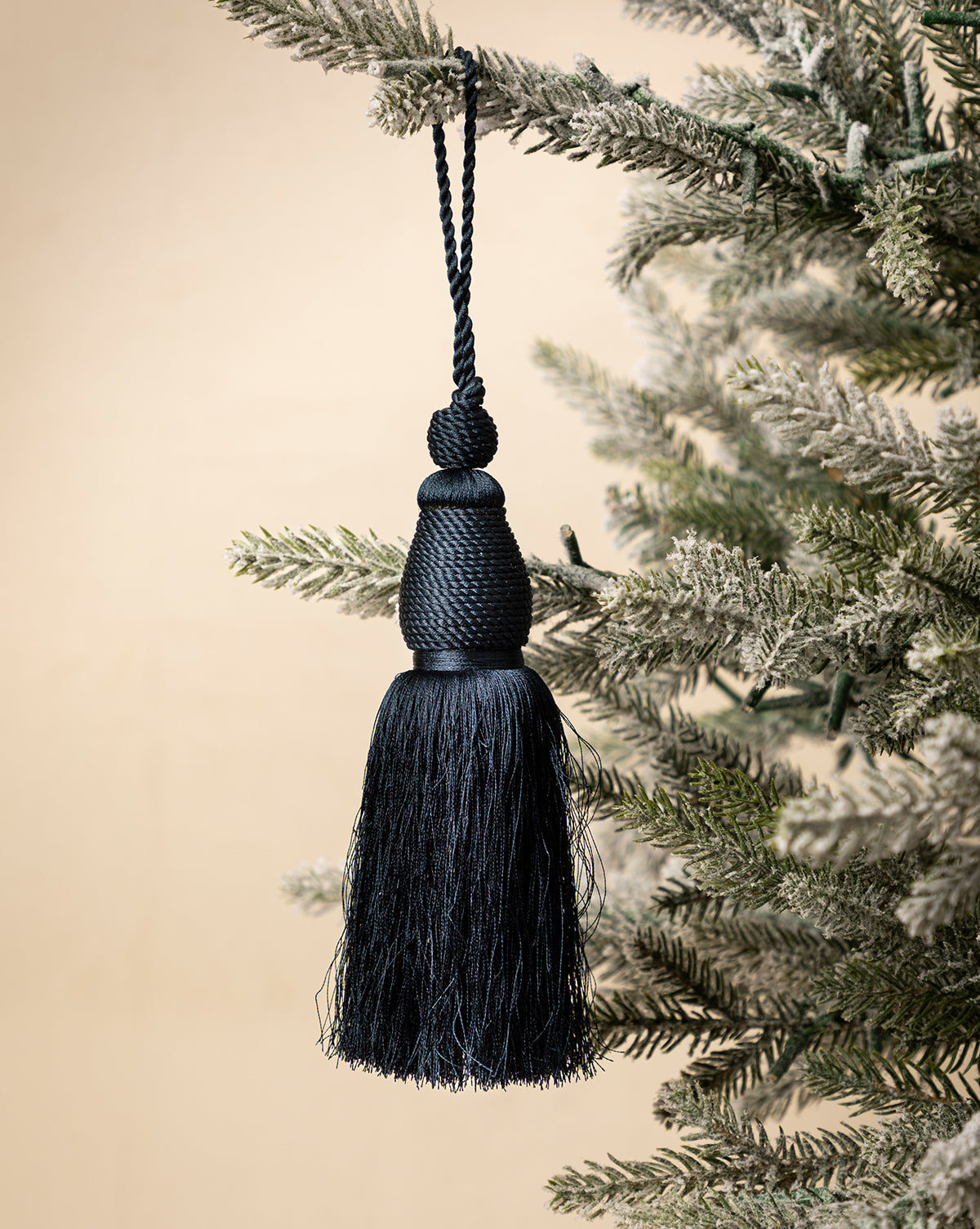 A Lucia Bullion Tassel in black brings vintage-inspired charm to your holiday décor, hanging as an ornament from a snow-frosted artificial evergreen branch against a neutral background, by Vendixs