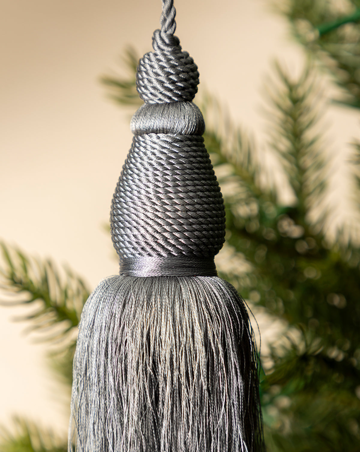 The Lucia Bullion Tassel, a vintage-inspired charm, is shown in close-up against blurred green pine branches and a neutral background, by Vendixs