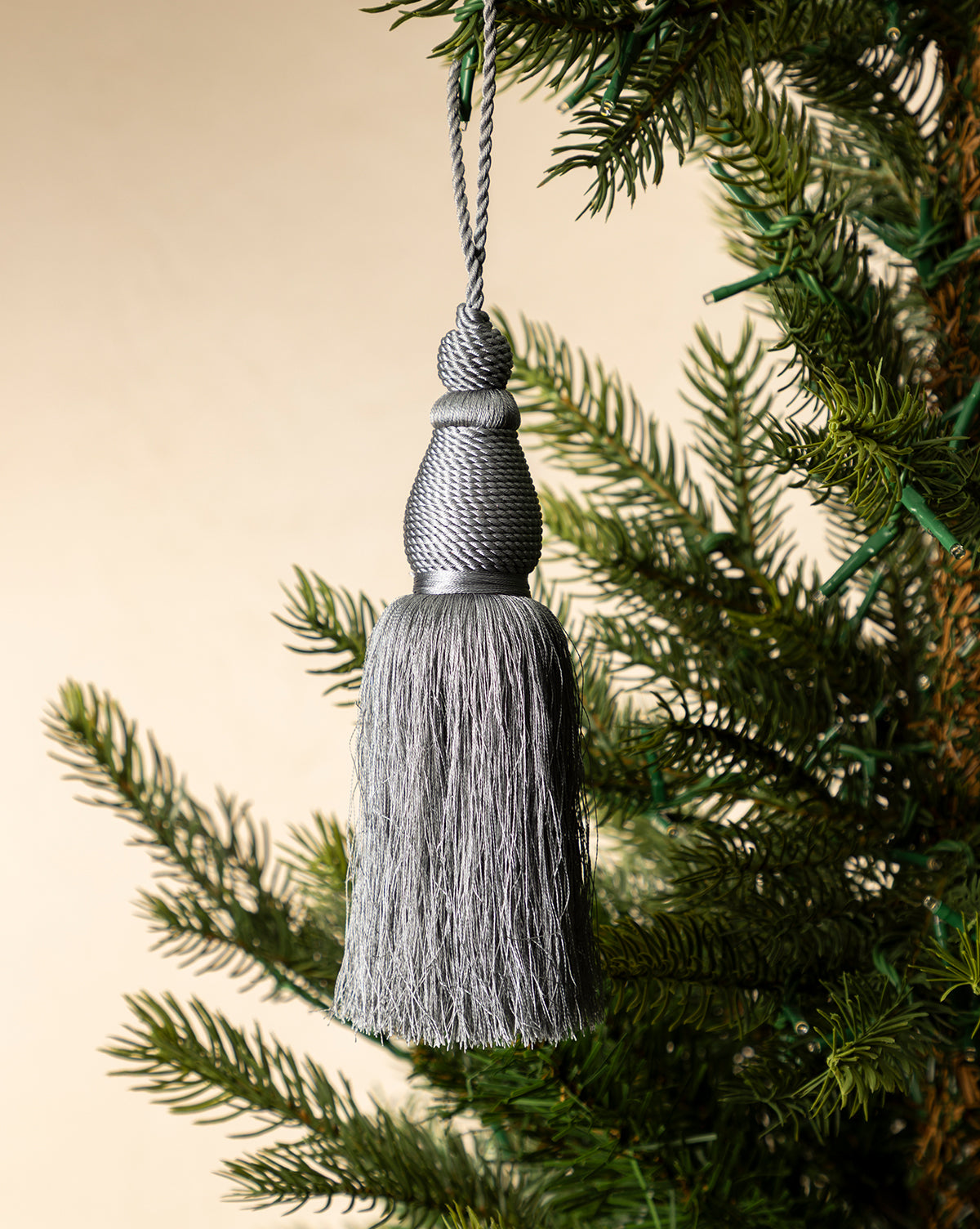 The Lucia Bullion Tassel hangs from a green pine branch, bringing vintage-inspired charm to a neutral beige background, by Vendixs
