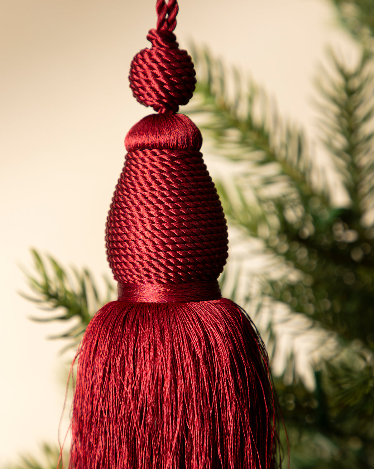 A close-up of the Lucia Bullion Tassel, a red holiday tree ornament, hanging with softly blurred green pine branches in the background, by Vendixs