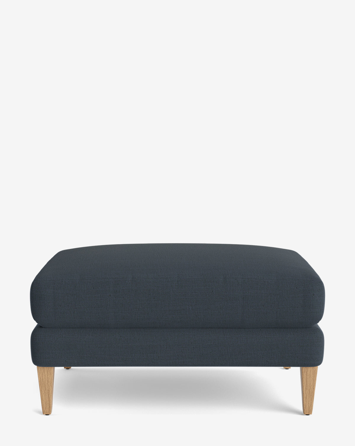The Lucille Ottoman is a modern, rectangular ottoman with dark blue upholstery and light wood legs, displayed on a plain white background by McGee & Co.