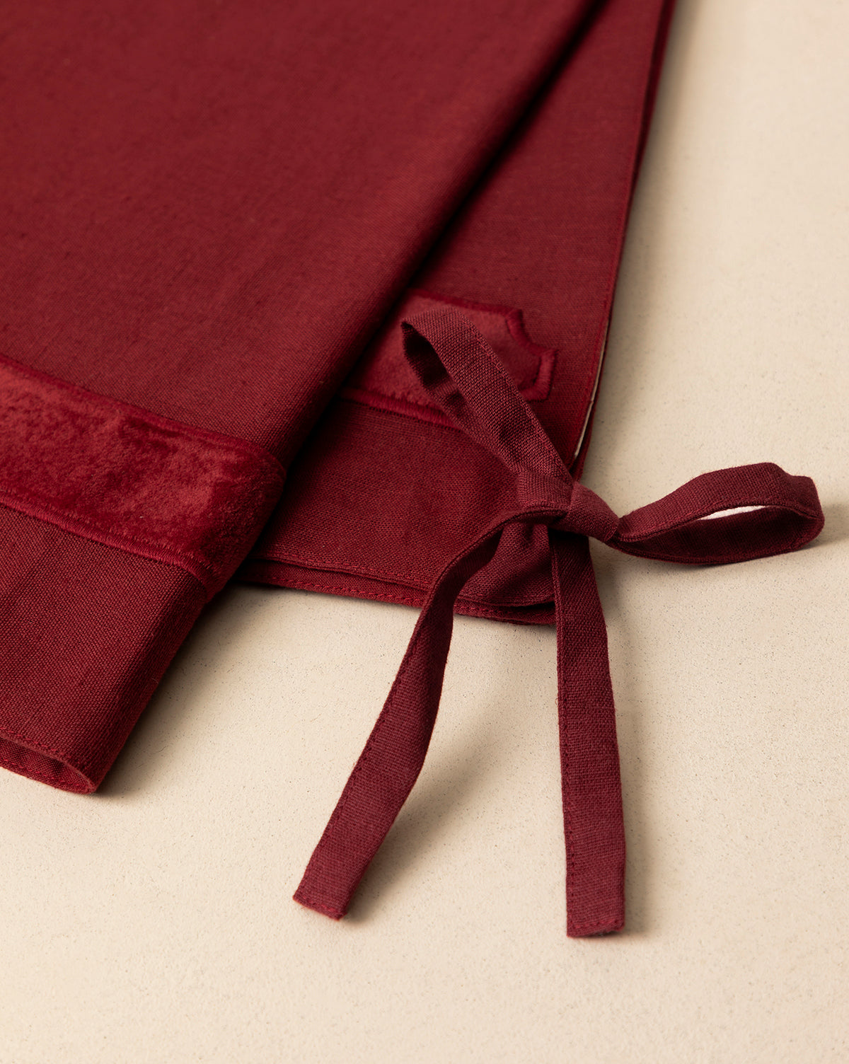 Close-up of the Mabry Velvet Trimmed Tree Skirt in deep red, folded on a light beige surface. The velvet texture and tie detail are clearly visible, by Vendixs