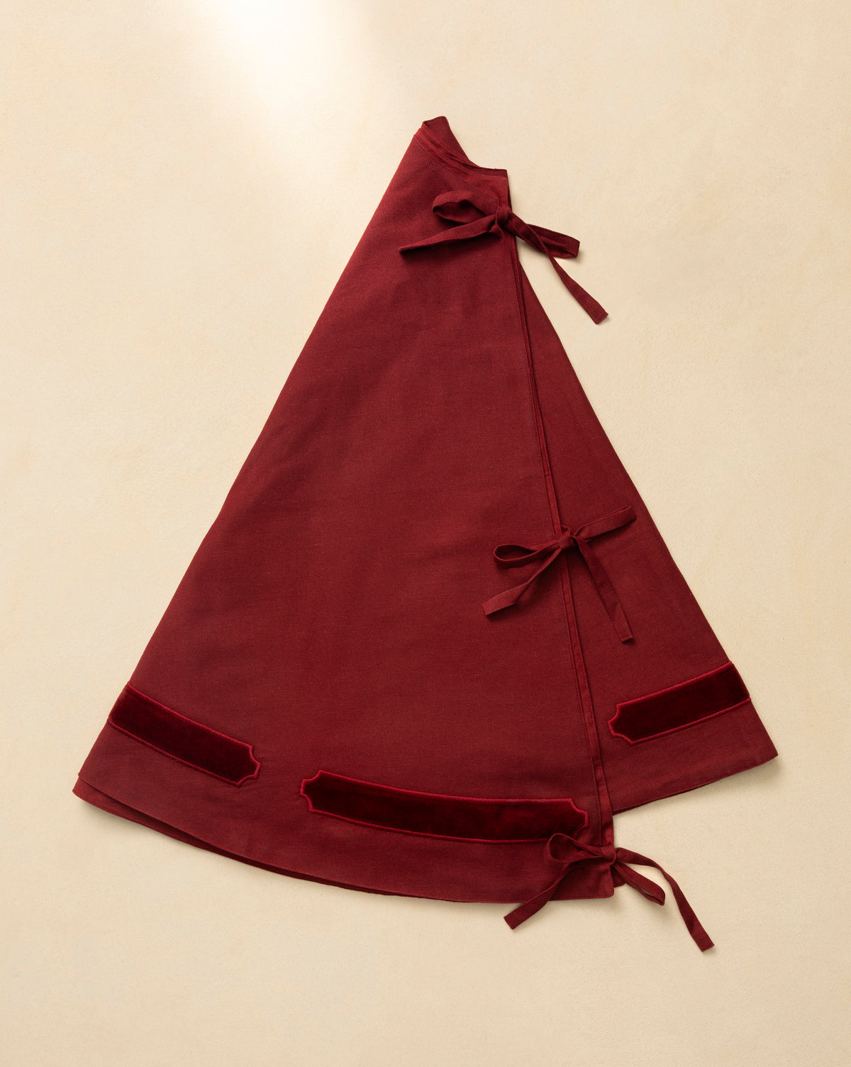 The Mabry Velvet Trimmed Tree Skirt is a deep red, triangular wrap skirt with three side fabric ties and two velvet trims near the hem, shown flat on beige—ideal for adding unique luxury to your holiday decor, by Vendixs