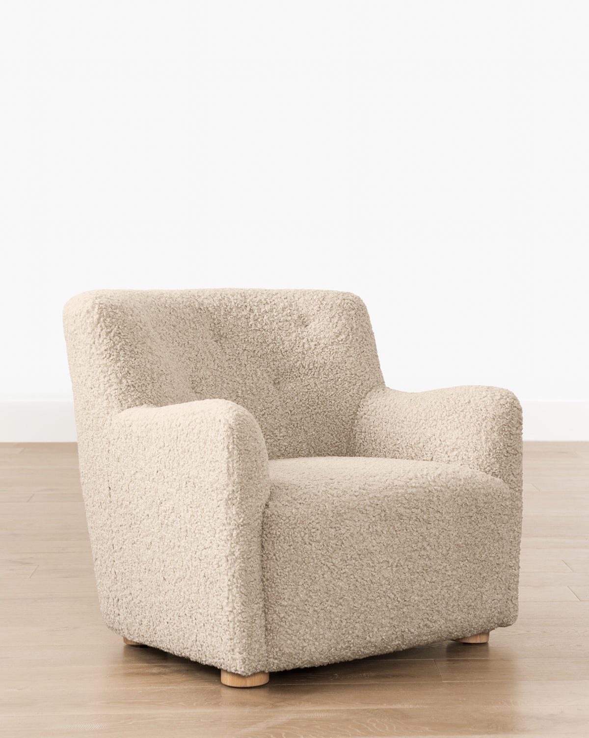 The Magda Lounge Chair (Ready to Ship) features plush, textured beige fabric, rounded arms, and short wooden legs. Its modern design adds a stylish touch when placed on a light wood floor against a white wall, by Vendixs