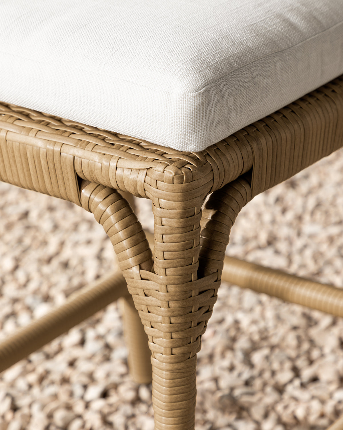 Close-up of the Makerspalm Marisol Indoor/Outdoor Counter Stool (Set of 2), showcasing a handwoven wicker leg and white cushioned seat, displayed on small beige pebbles. Vendixs