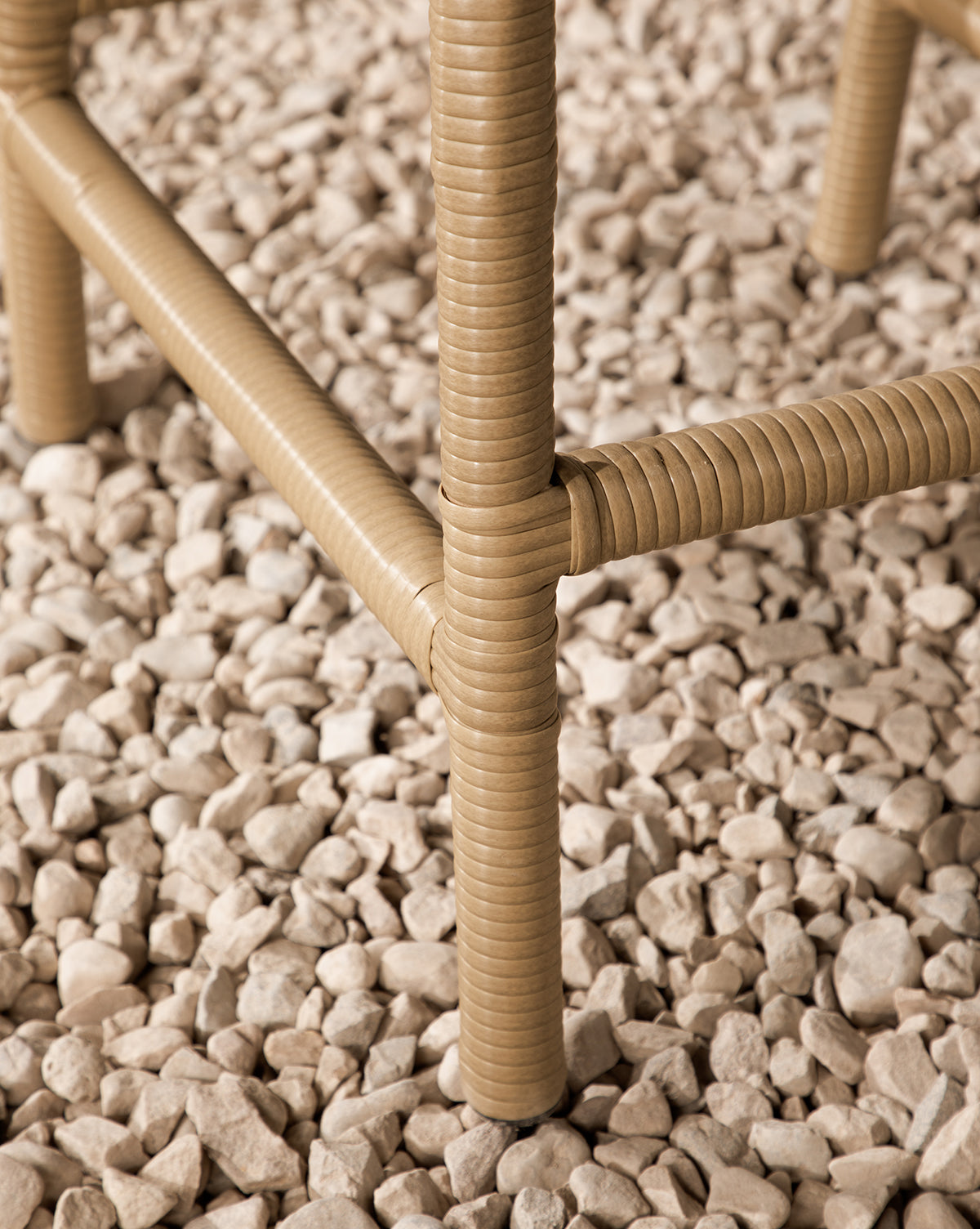 Close-up of tan, textured legs from the Makerspalm Marisol Indoor/Outdoor Counter Stool (Set of 2), standing on a bed of small, light beige pebbles. Vendixs