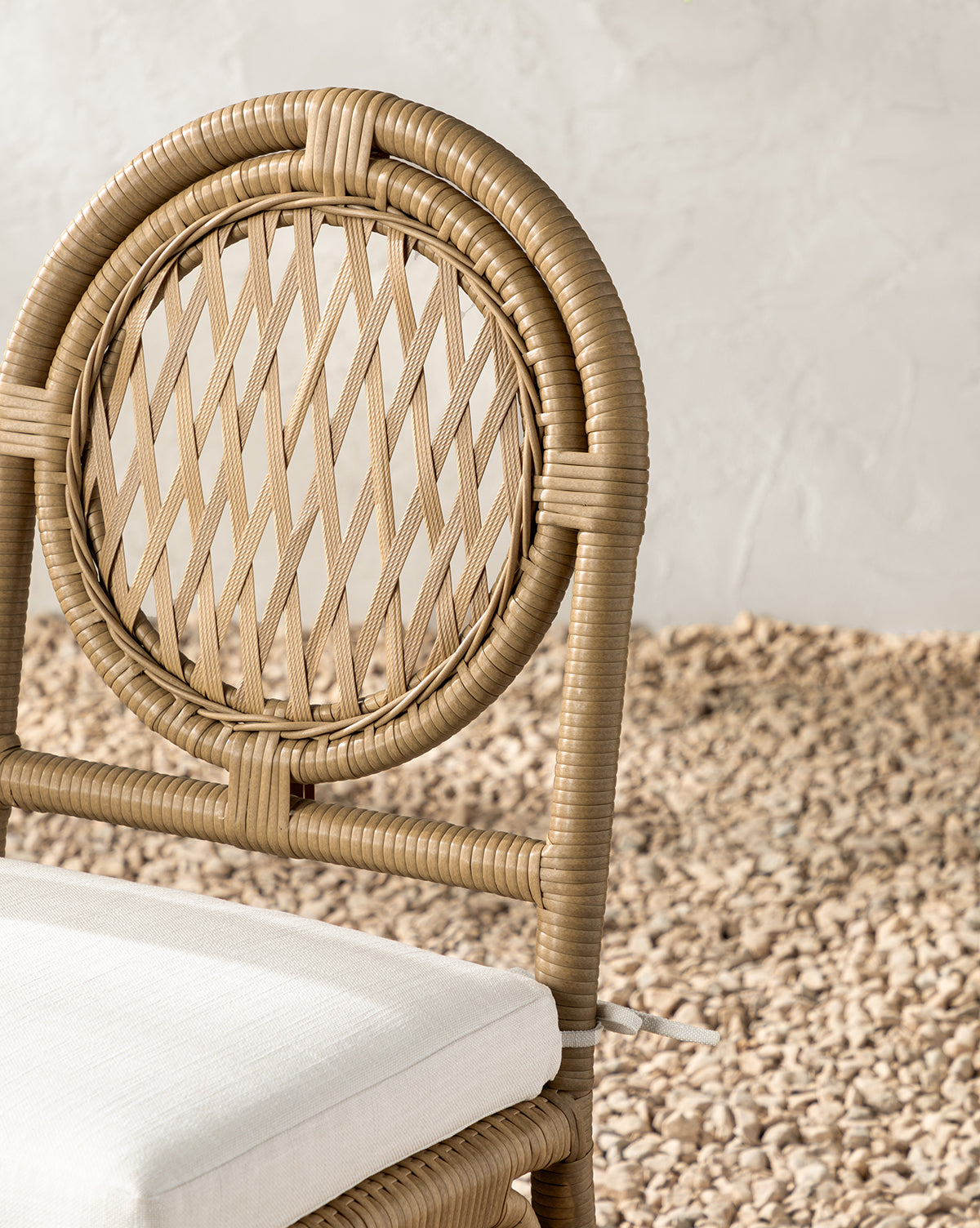 A close-up of the Marisol Indoor/Outdoor Dining Chair (Set of 2), featuring a weather-resistant, round handwoven wicker backrest and white cushioned seat, set on pebbles against a textured white wall, by Vendixs