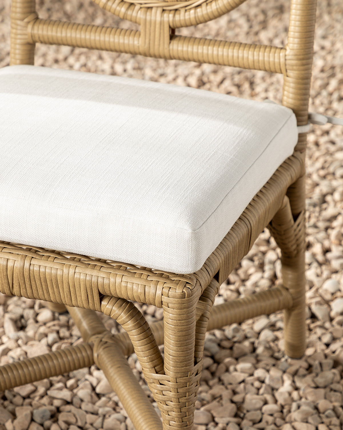 A close-up of the Marisol Indoor/Outdoor Dining Chair (Set of 2) featuring handwoven wicker and a white cushioned seat, placed outside on small beige pebbles, by Vendixs
