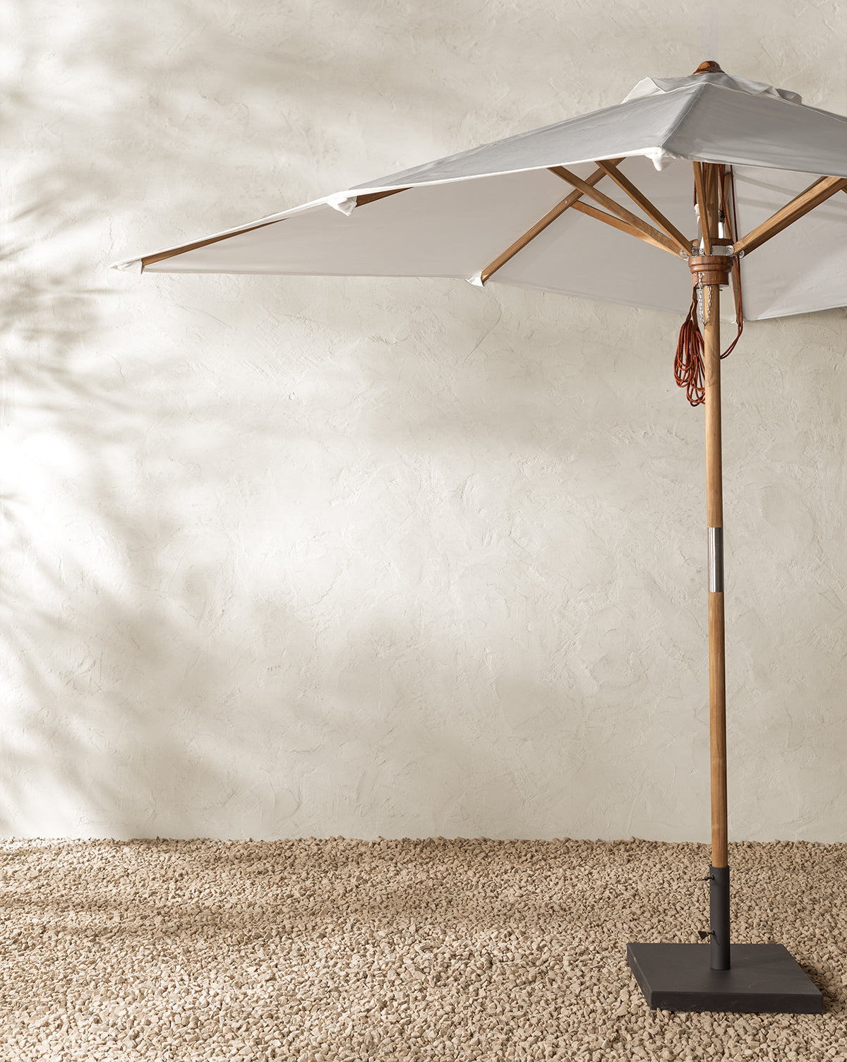 The Marques Outdoor Rectangular Umbrella, featuring a large white canopy and teak pole, stands on a textured gravel surface against a light beige stucco wall, casting soft shadows in this minimalist outdoor space, by McGee & Co.