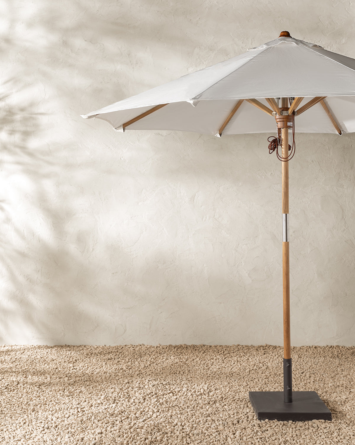 The Marques Outdoor Round Umbrella features a weather-resistant acrylic white canopy and sturdy teak pole, standing on textured gravel against a light beige stucco wall with soft background shadows, by McGee & Co.
