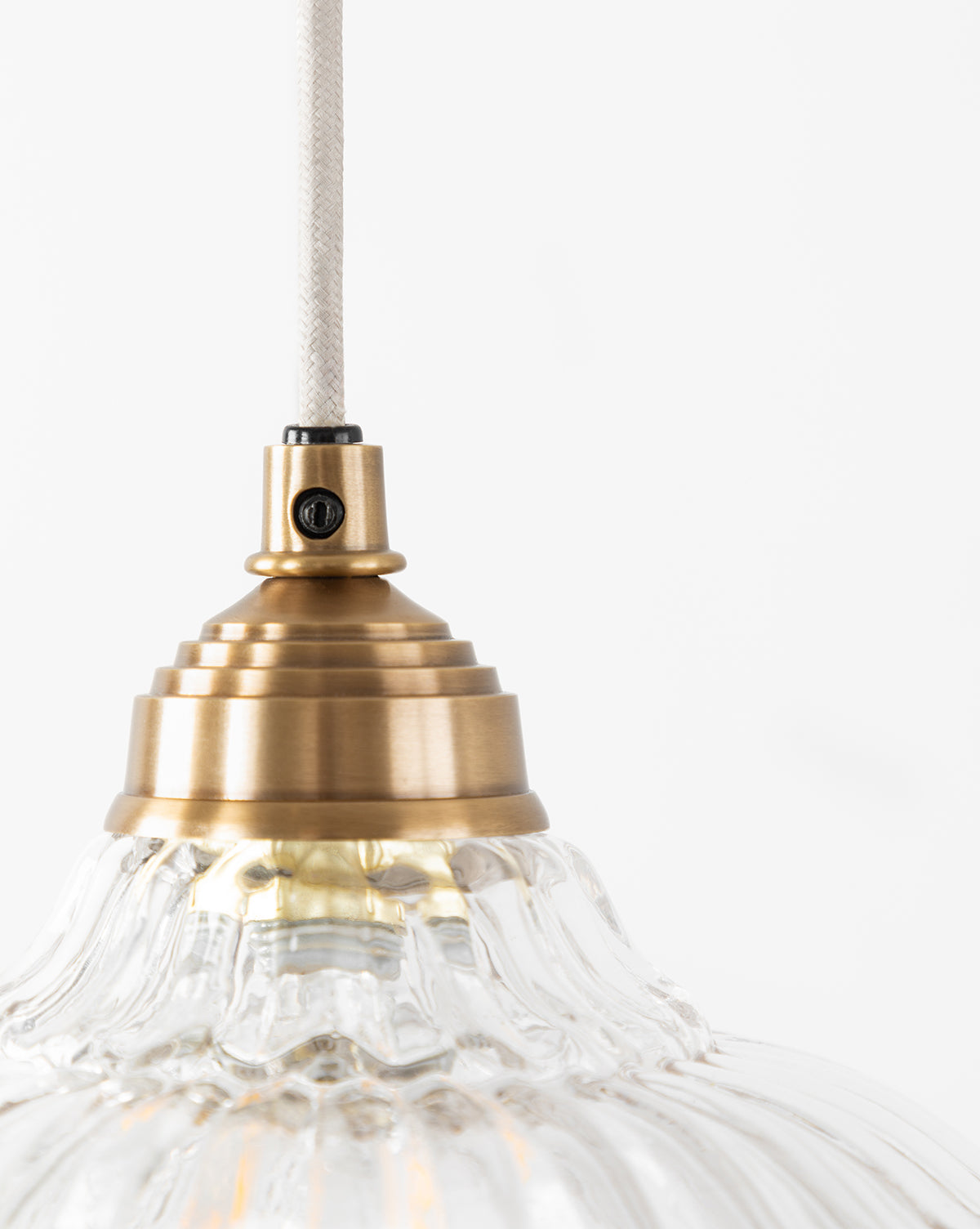A close-up of the Vendixs x Loeffler Randall Maude Glass Pendant shows its ribbed clear glass shade and brass hardware, suspended by a white cord against a plain white background, by Vendixs