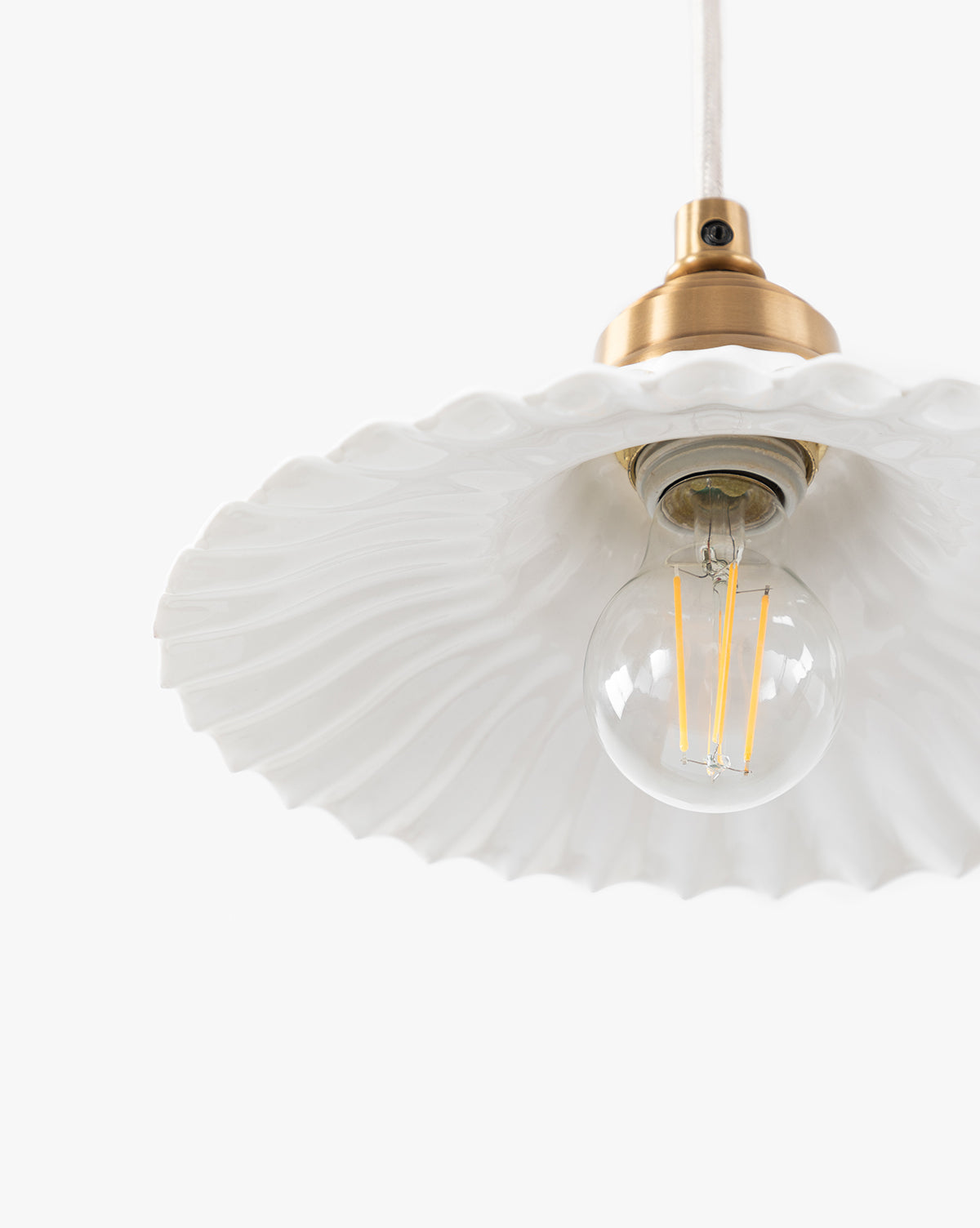 A close-up of the Vendixs x Loeffler Randall Maude Glass Pendant shows brass hardware, a clear round bulb, and a white wavy-edged ceramic shade, set against a plain white background by Vendixs