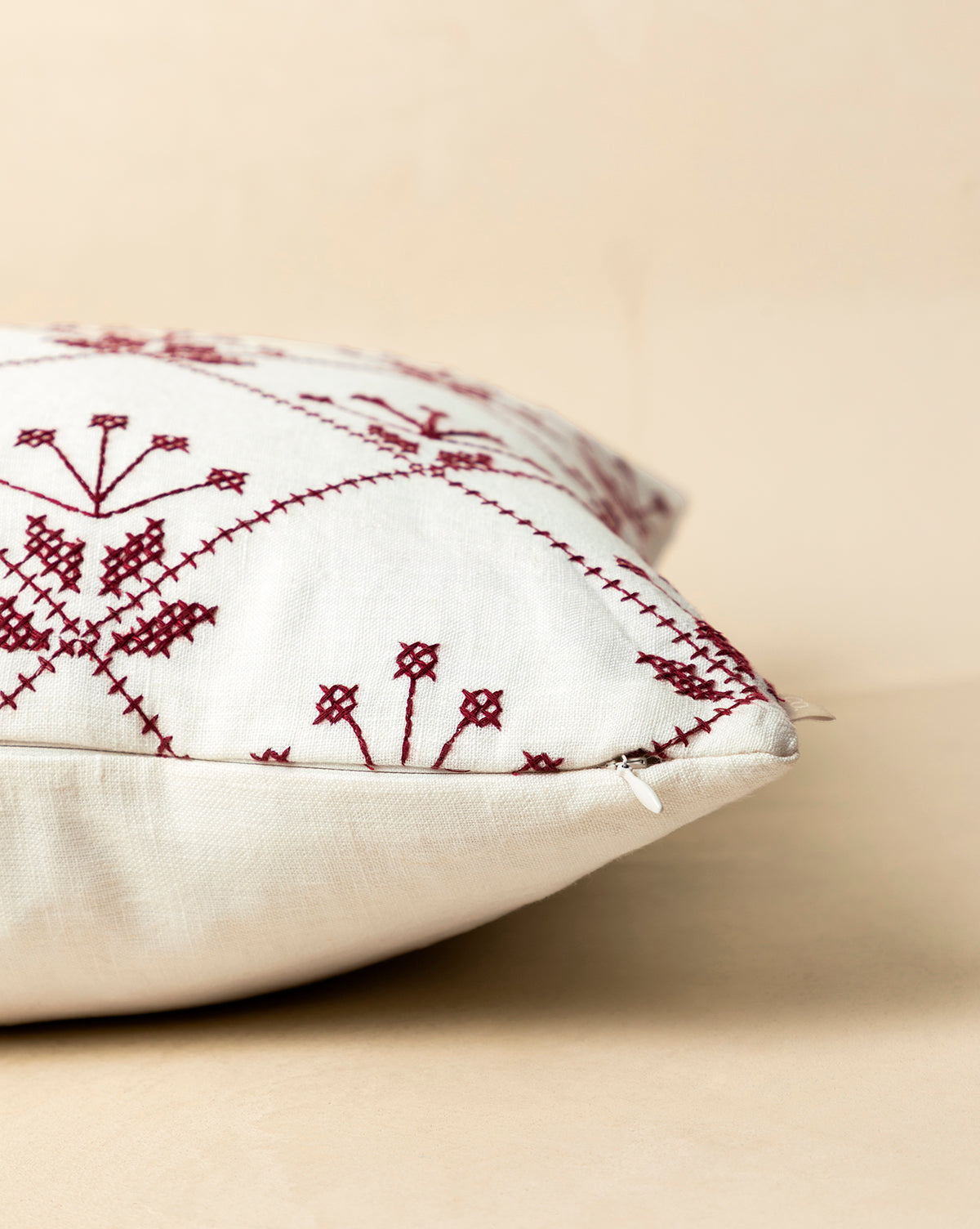 A close-up of the McCallister Pillow Cover showcases maroon embroidered geometric and floral designs on white fabric, set on a beige background. A subtle zipper adds elegance—perfect for holiday home decor, by Vendixs