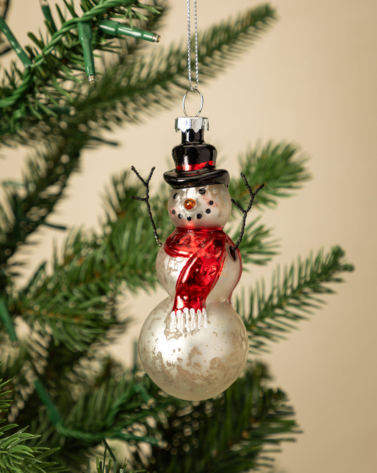 The Mercury Glass & Wire Snowman Ornament, featuring a red scarf, black top hat, and twig arms, hangs from a green Christmas tree branch, bringing vintage charm to your holiday décor against a warm beige background, by McGee & Co.