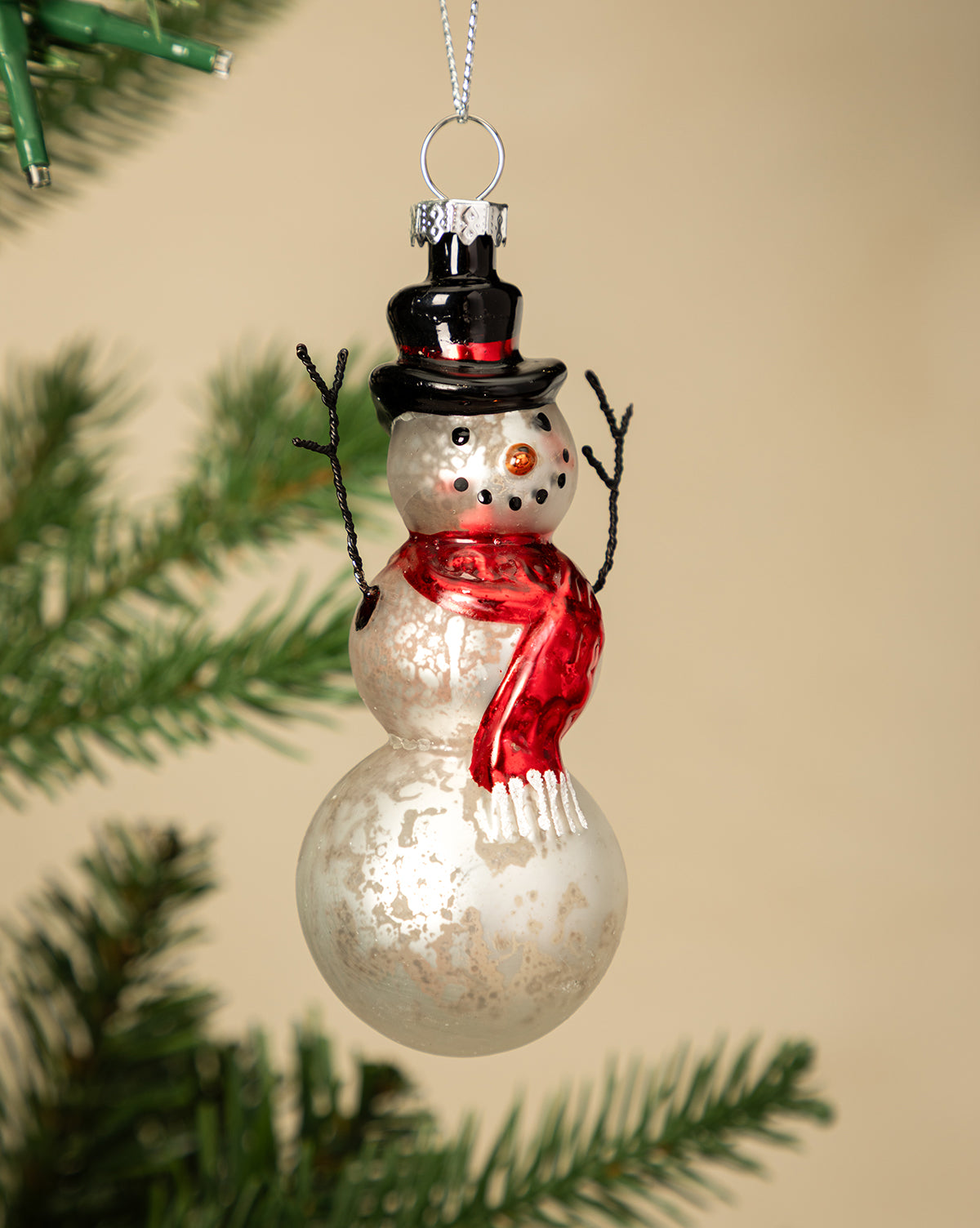 The Mercury Glass & Wire Snowman Ornament, featuring a black top hat, red scarf, and stick arms, hangs from a Christmas tree branch against a beige background, bringing vintage-inspired charm to your holiday decor, by Vendixs