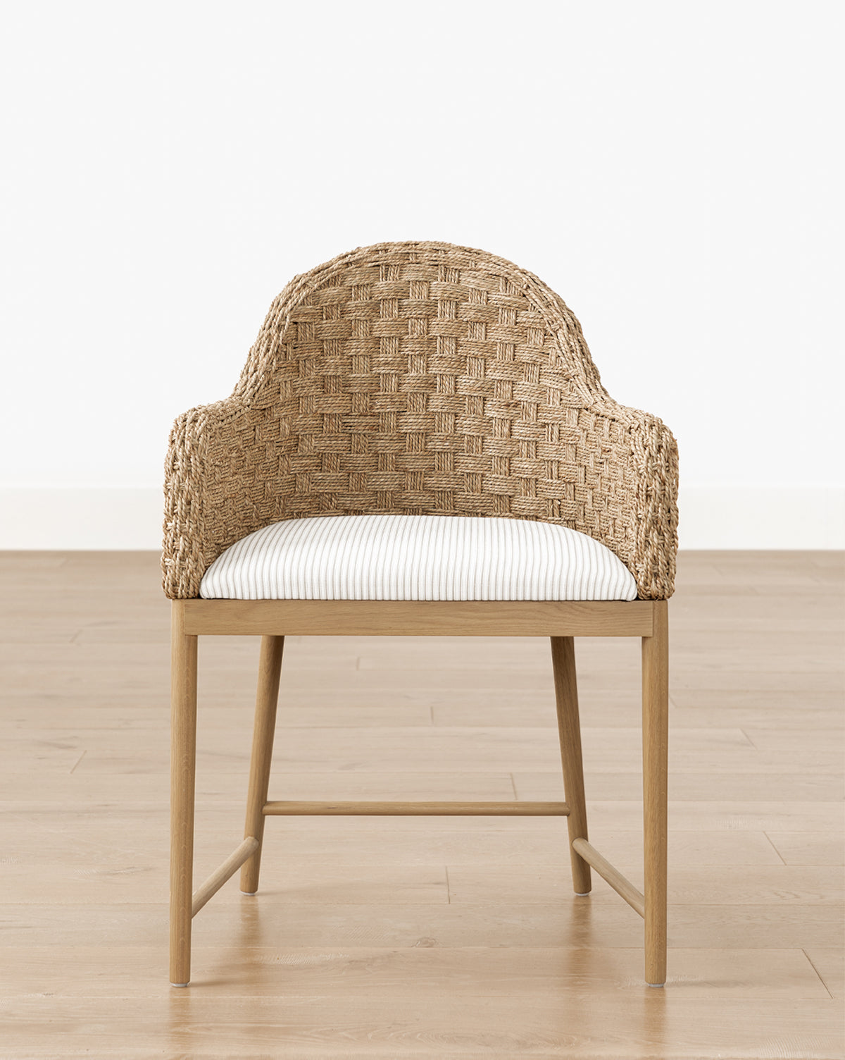 The Makerspalm Molly Arm Chair features woven rattan, a curved back, white striped cushion, and solid oak legs, shown on a light wood floor against a plain white wall. Vendixs