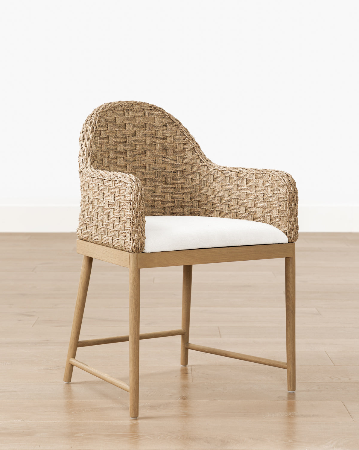 The Molly Arm Chair features a modern woven rattan design with curved back and arms, a light wood frame, and a white cushioned seat, set on a light wood floor against a plain white wall by Vendixs