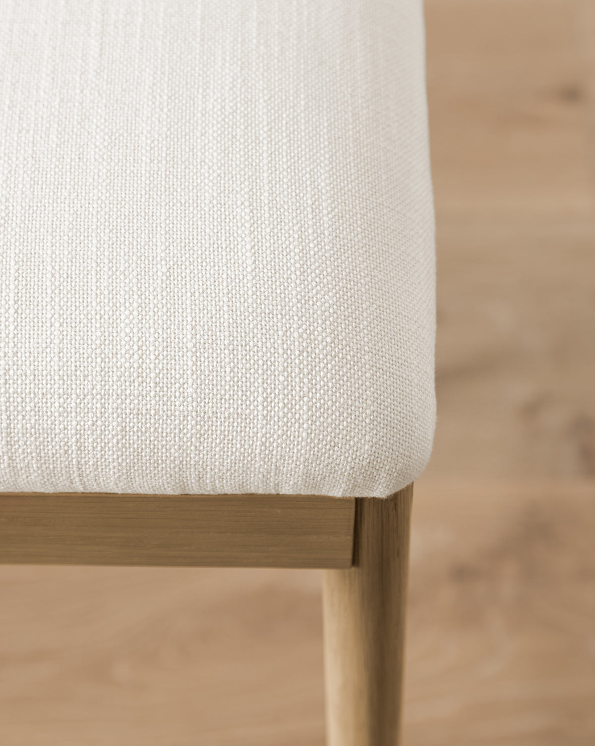 A close-up of the Molly Dining Chair shows its light beige, textured upholstery and solid oak frame with wooden legs, set on a light wood floor. The image focuses on the corner to highlight the detailed fabric and craftsmanship by McGee & Co.
