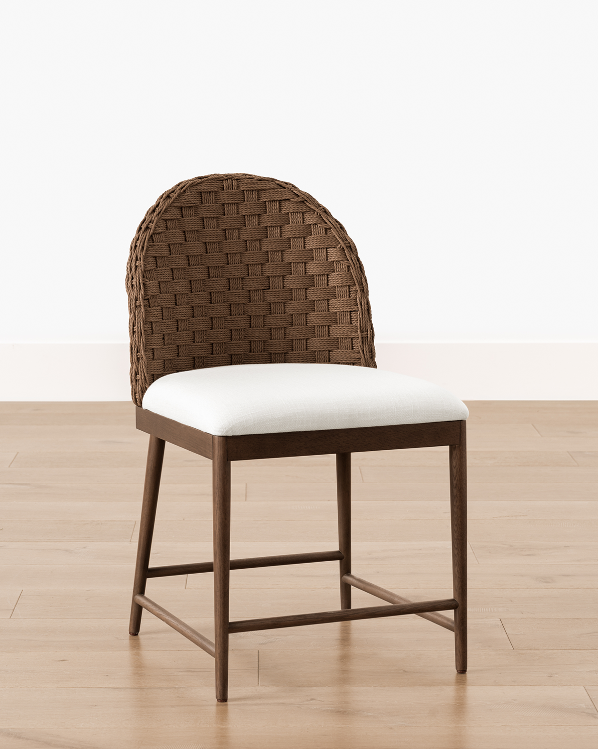 The Molly Dining Chair showcases a wooden frame, a woven rounded backrest, and a white cushioned seat, elegantly set on a light wooden floor by a white wall, by Vendixs