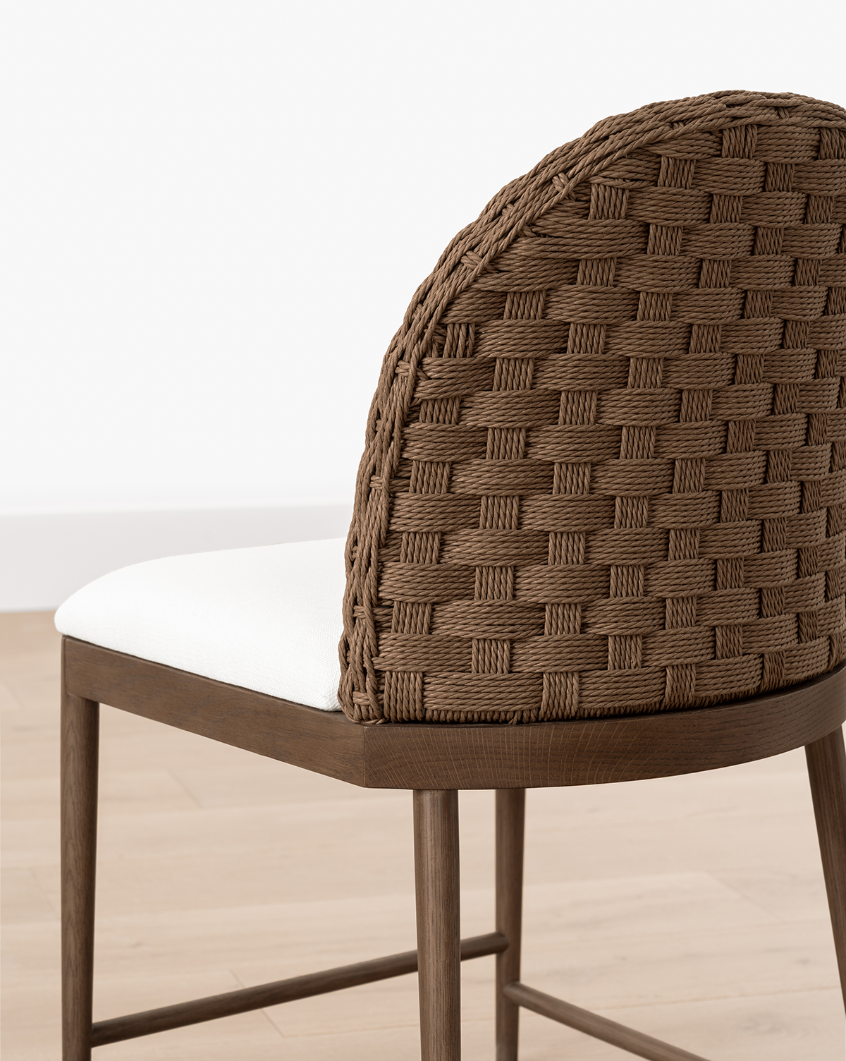 The Molly Dining Chair features a woven brown backrest, white cushioned seat, and sturdy wooden legs, shown here on a light wood floor against a white wall, by Vendixs