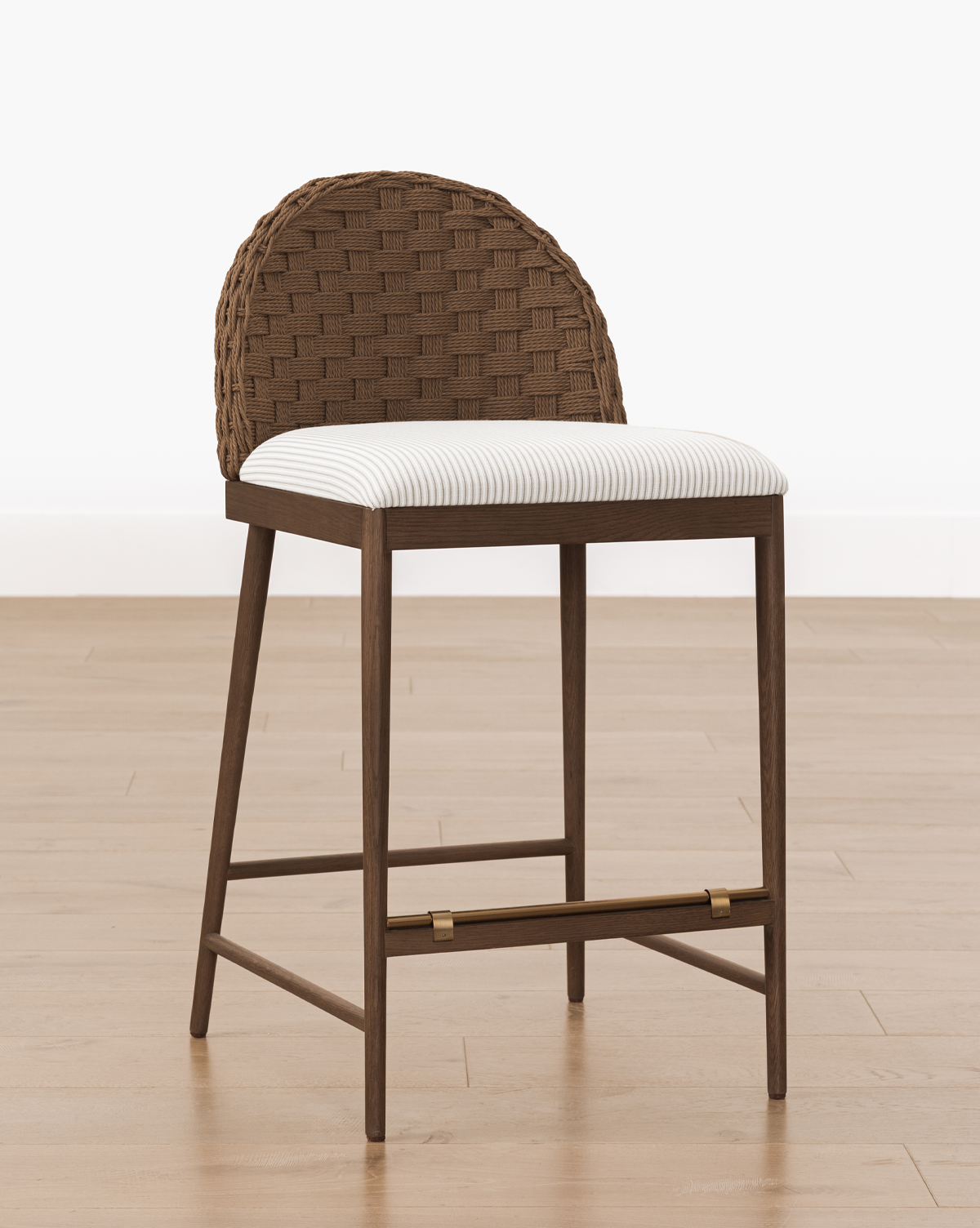 The Molly Stool features a natural finish, a woven rounded backrest, and a cushioned seat in light striped fabric, shown on a light wood floor against a plain white wall, by Vendixs