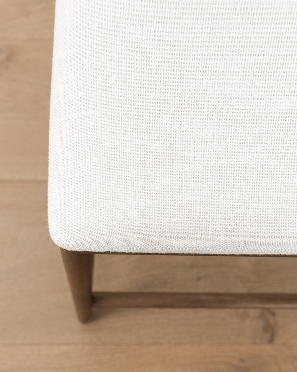 A detailed view of the Molly Stool’s corner highlights its solid oak frame and textured off-white fabric seat, set atop a light wooden floor, by Vendixs