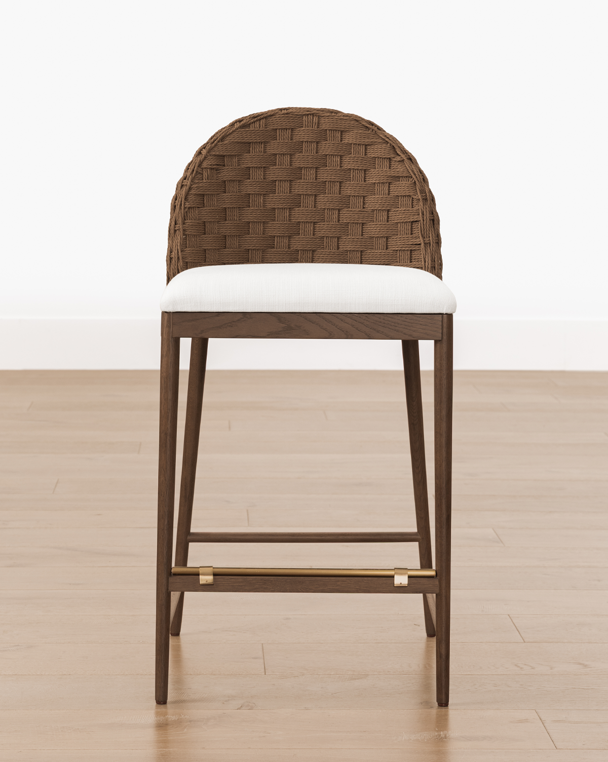The Molly Stool features a woven brown backrest, white cushioned seat, and solid oak legs with a brass footrest, shown on a light wooden floor against a white wall, by Vendixs