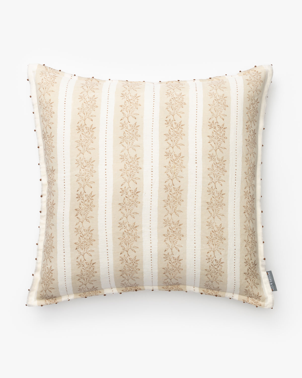 The Nettles Pillow Cover is a square linen cover with cream and beige vertical stripes, a subtle floral pattern, small decorative beads on the edges, and a fabric tag on the right side. Shown against a plain white background by Vendixs