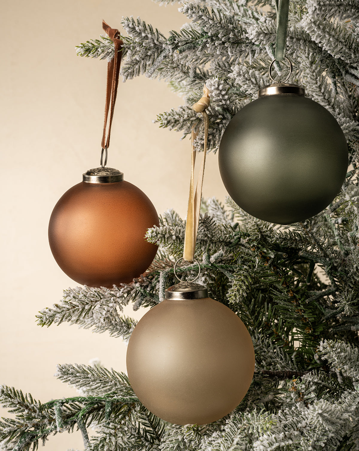 Three frosted Noel Festive Ornaments from the 9-piece set—brown, green, and gold glass—hang on a snow-dusted artificial Christmas tree against a neutral background, by Vendixs