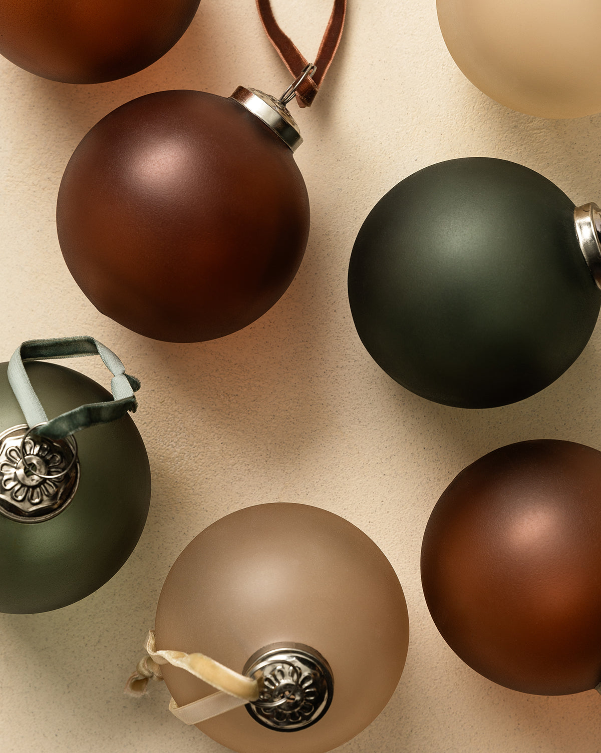 A flat lay features the Noel Festive Ornaments (Set of 9) in matte glass with earthy brown, green, and beige tones, each with decorative silver caps and assorted ribbon hangers, arranged on a neutral background for Christmas décor, by Vendixs