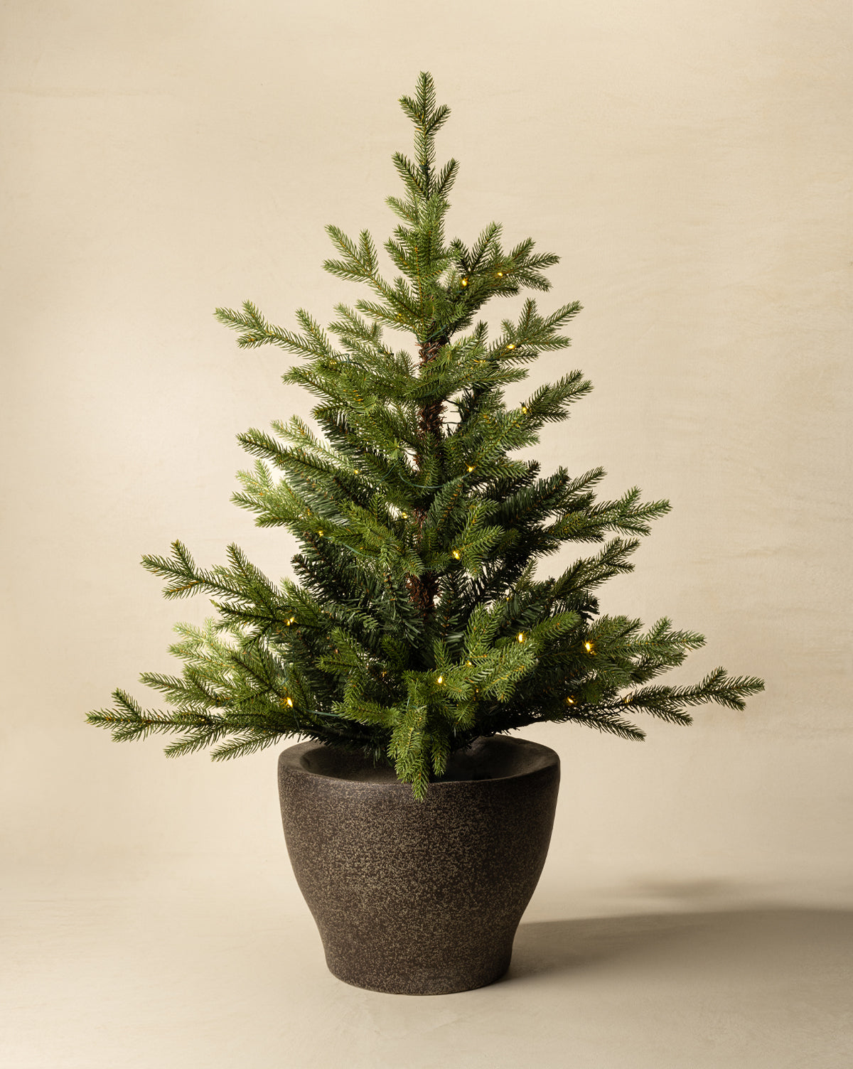 The 3' Norway Pre-Lit Faux Potted Tree features warm white lights and sits in a dark, textured ceramic pot, set against a plain beige background, by Vendixs