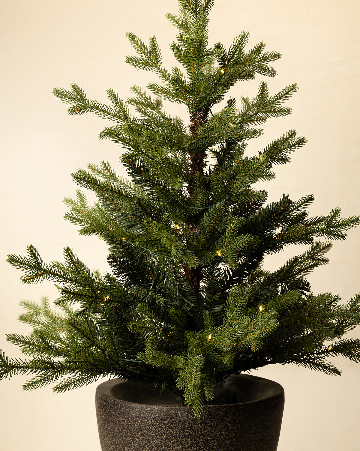 The 3' Norway Pre-Lit Faux Potted Tree features sparsely placed branches and warm white lights in a dark, round ceramic pot, set against a plain, light-colored background, by Vendixs