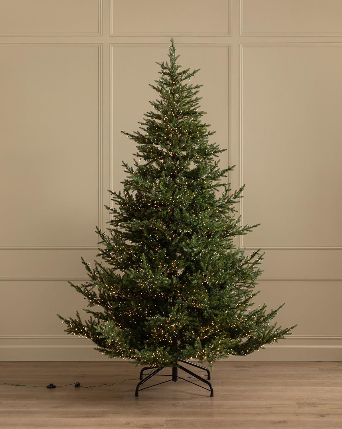 The 9' Norway Spruce Pre-Lit Faux Tree with warm white lights stands on a black metal stand in a room with beige paneled walls and a wooden floor, by Vendixs